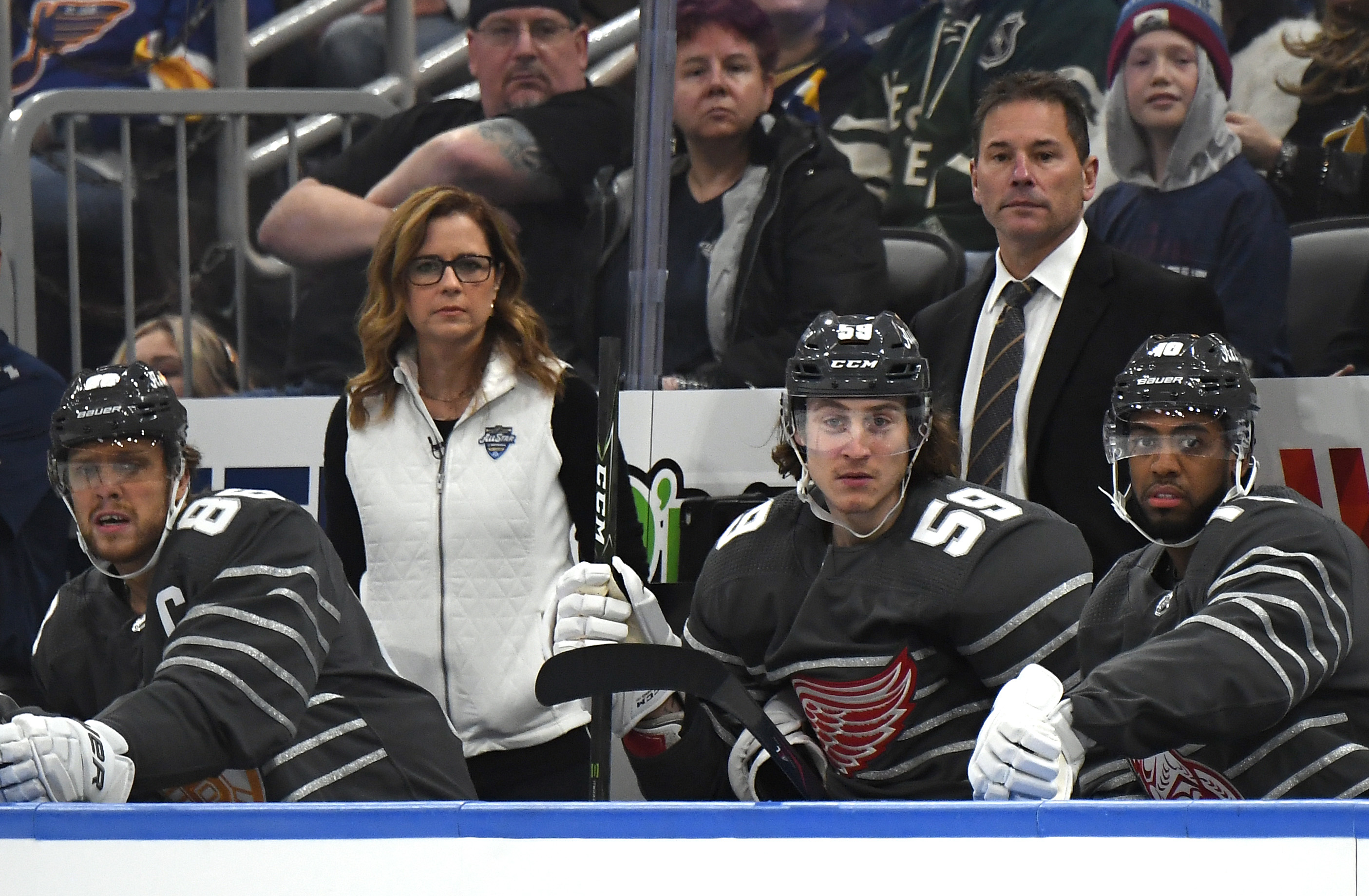 Several hockey players in uniform sit on the bench, with coaches and spectators in the background