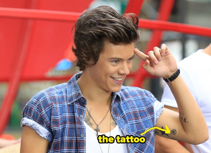 Harry Styles in a casual outfit with rolled-up sleeves, smiling and saluting with two fingers