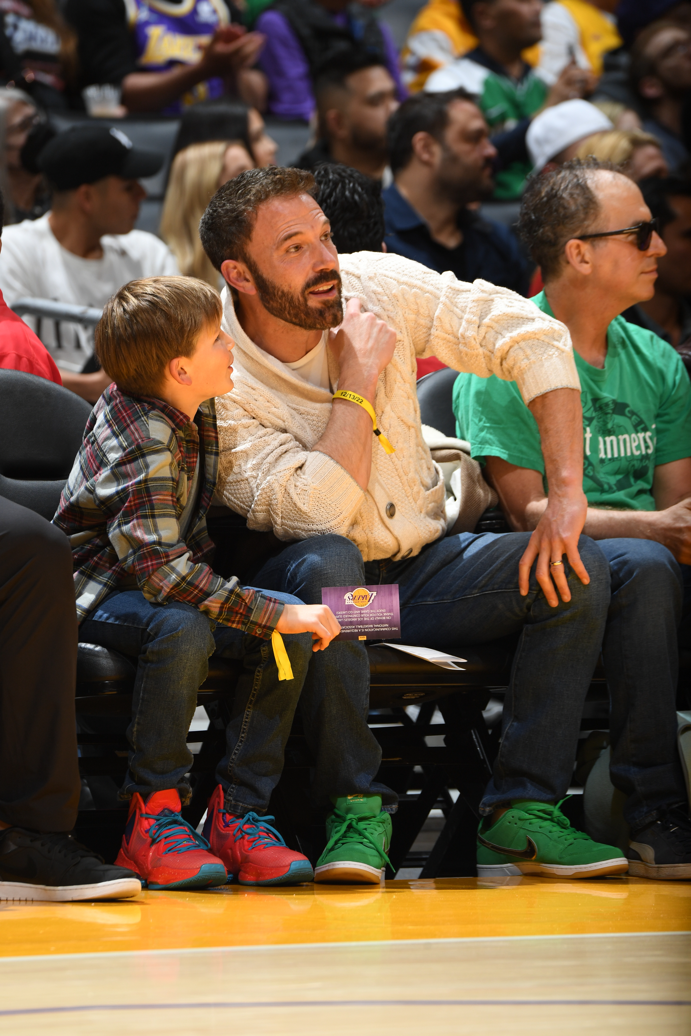 Ben Affleck sits courtside with his son, Samuel Affleck, at a basketball game, engaging in conversation and pointing towards the court