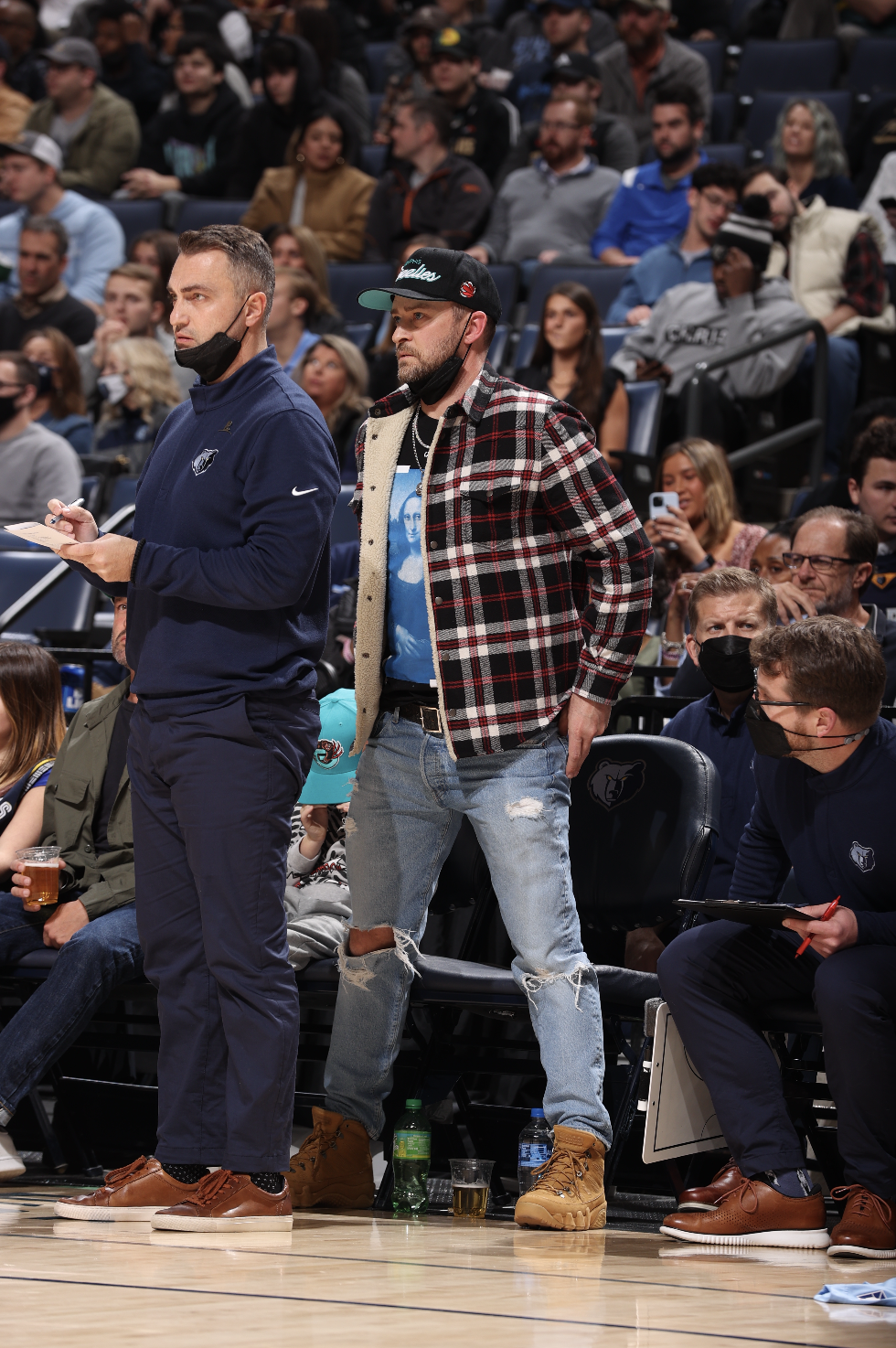 A man in a checkered jacket and ripped jeans stands courtside at a basketball game next to a man in a blue jacket and jeans. Fans are seated in the background