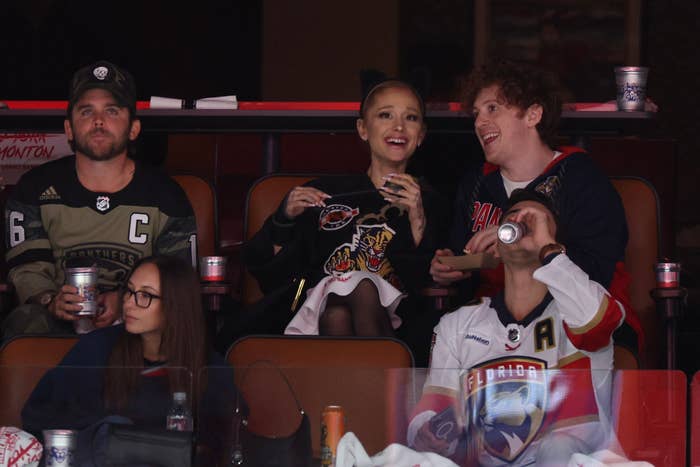 NHL game audience: Ariana Grande in a sweater, glasses and a hat, watches the game with Ethan Slater and other friends. Ethan wears a hockey jersey