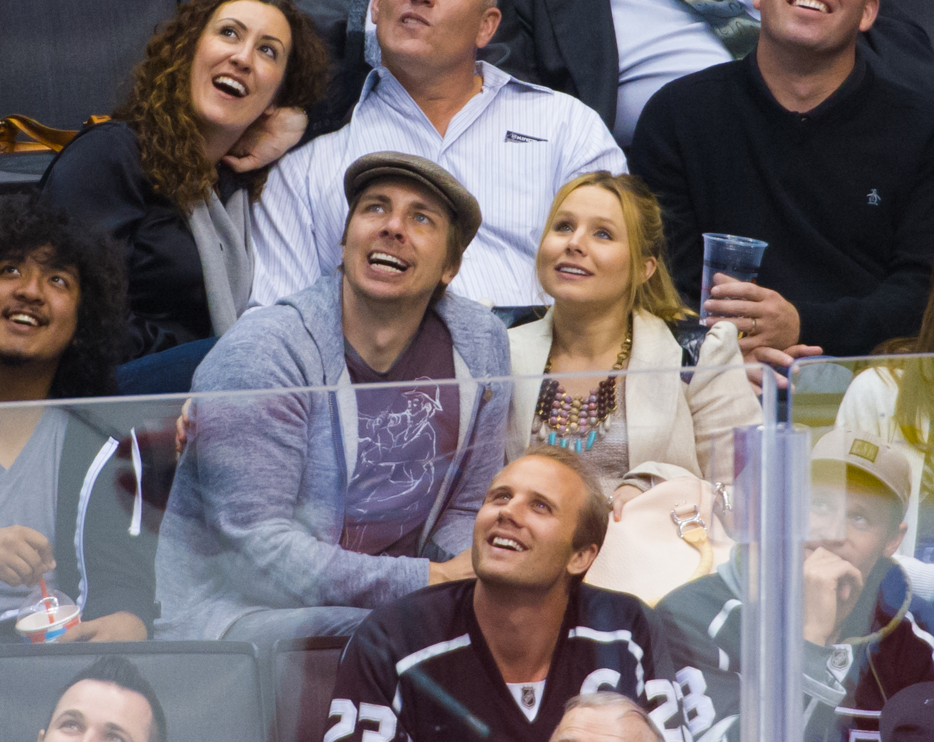 Dax Shepard, Kristen Bell, and others seated at a sporting event, looking upwards. Kristen is wearing a statement necklace, and Dax is in a casual outfit with a cap
