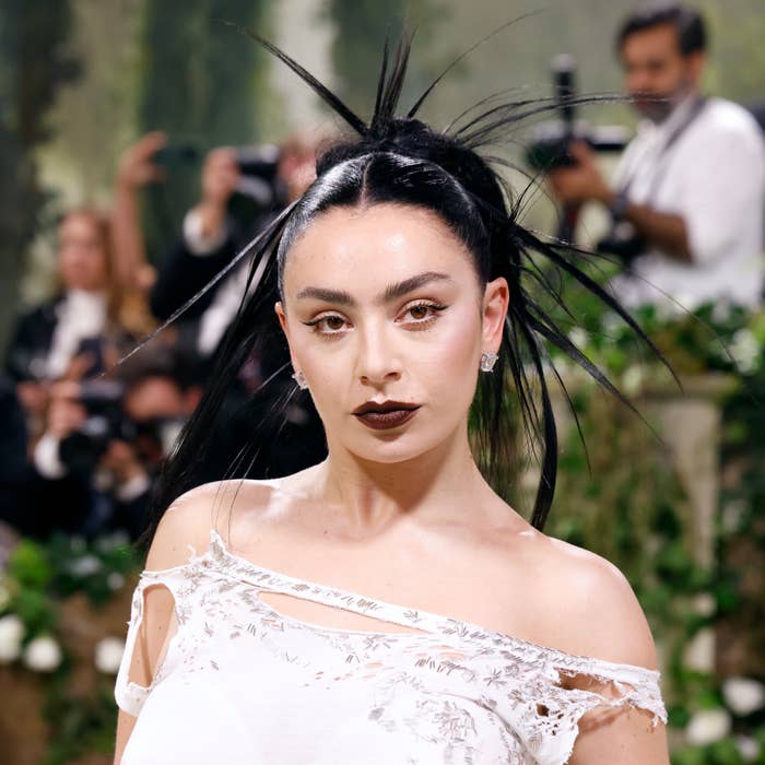 Charli XCX in an off-shoulder outfit on a red carpet, with photographers in the background. She has dramatic eye makeup and her hair is styled in a spiked updo