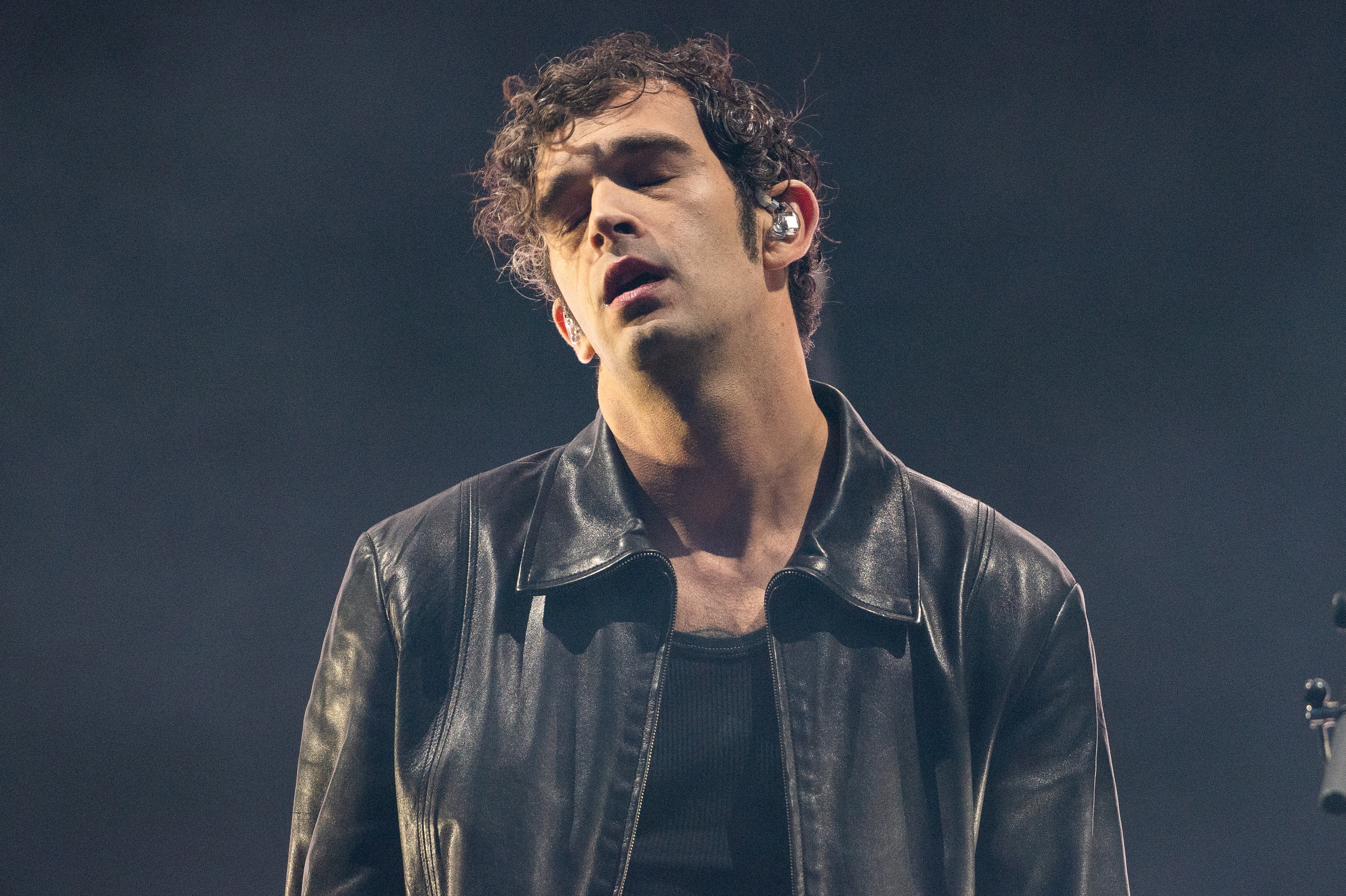 Matty Healy wearing a leather jacket and dark shirt, appearing to be in the middle of a performance with eyes closed and a focused expression