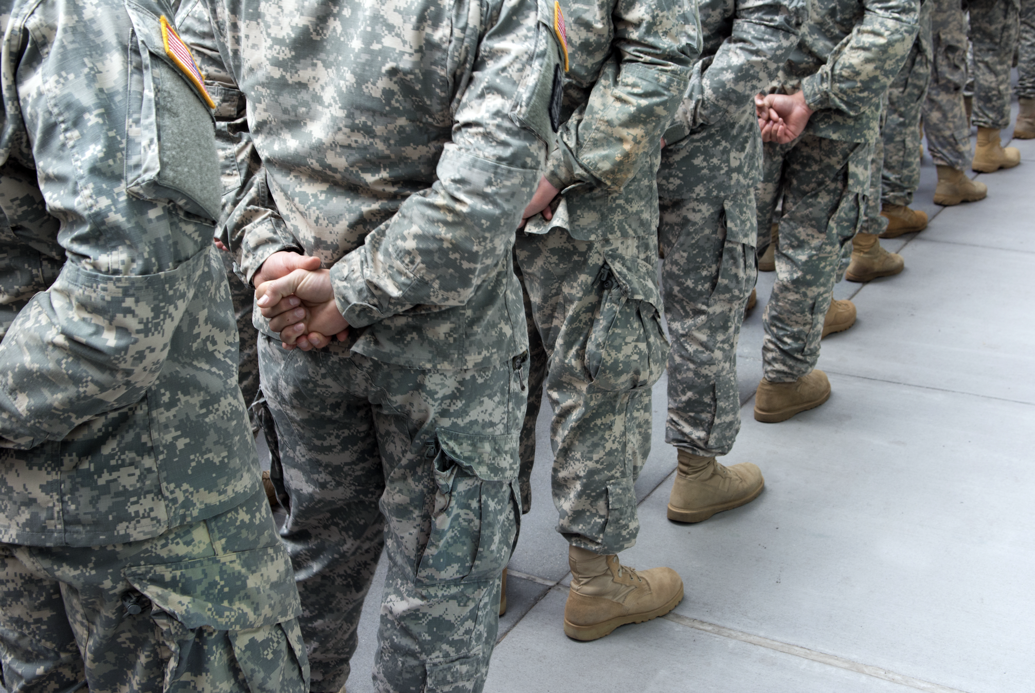 Soldiers in camouflage uniforms stand in a line with hands clasped behind their backs. The image focuses on the lower halves of their bodies