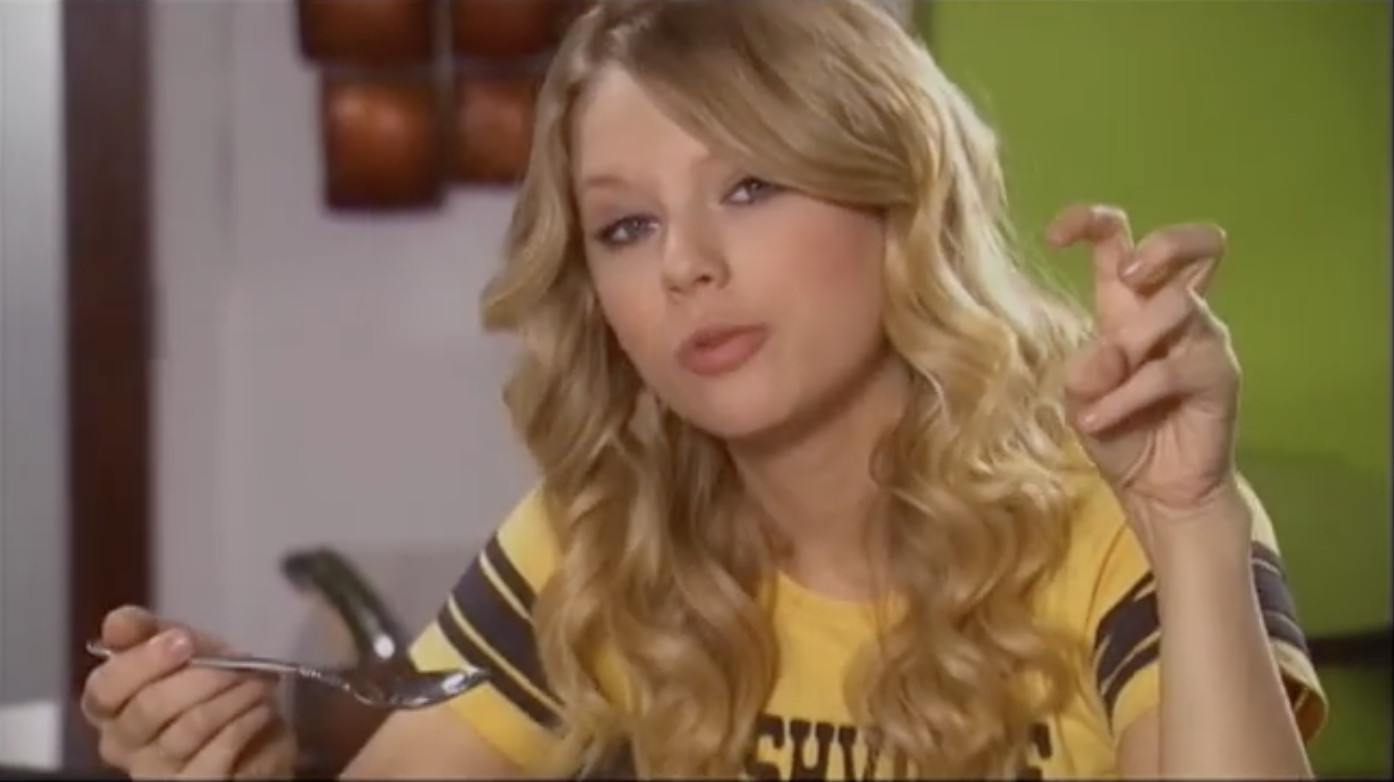 Taylor Swift is holding a spoon in one hand and making an air quote gesture with the other while speaking