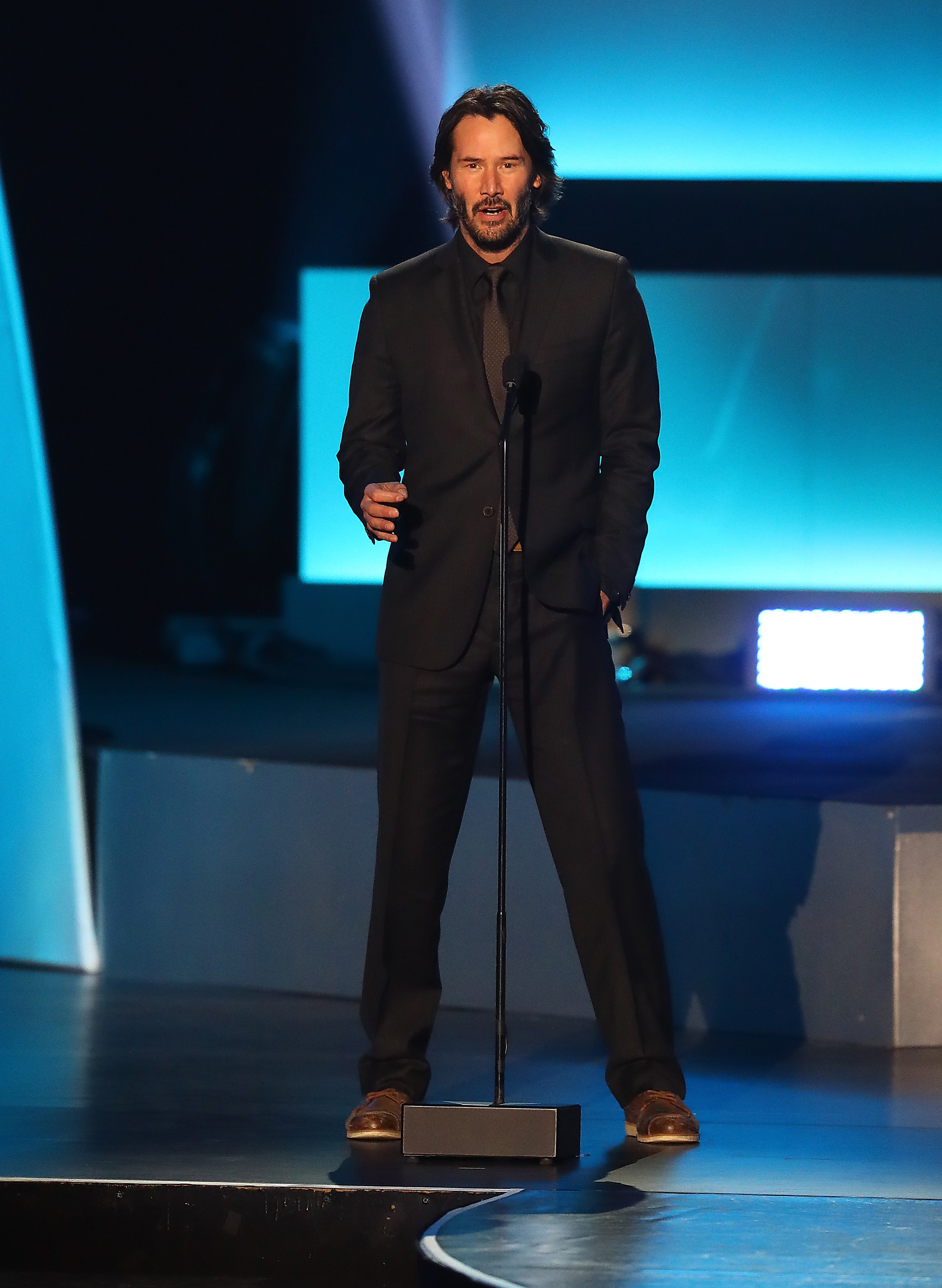 Keanu Reeves standing on stage in a black suit with a microphone, speaking at an event