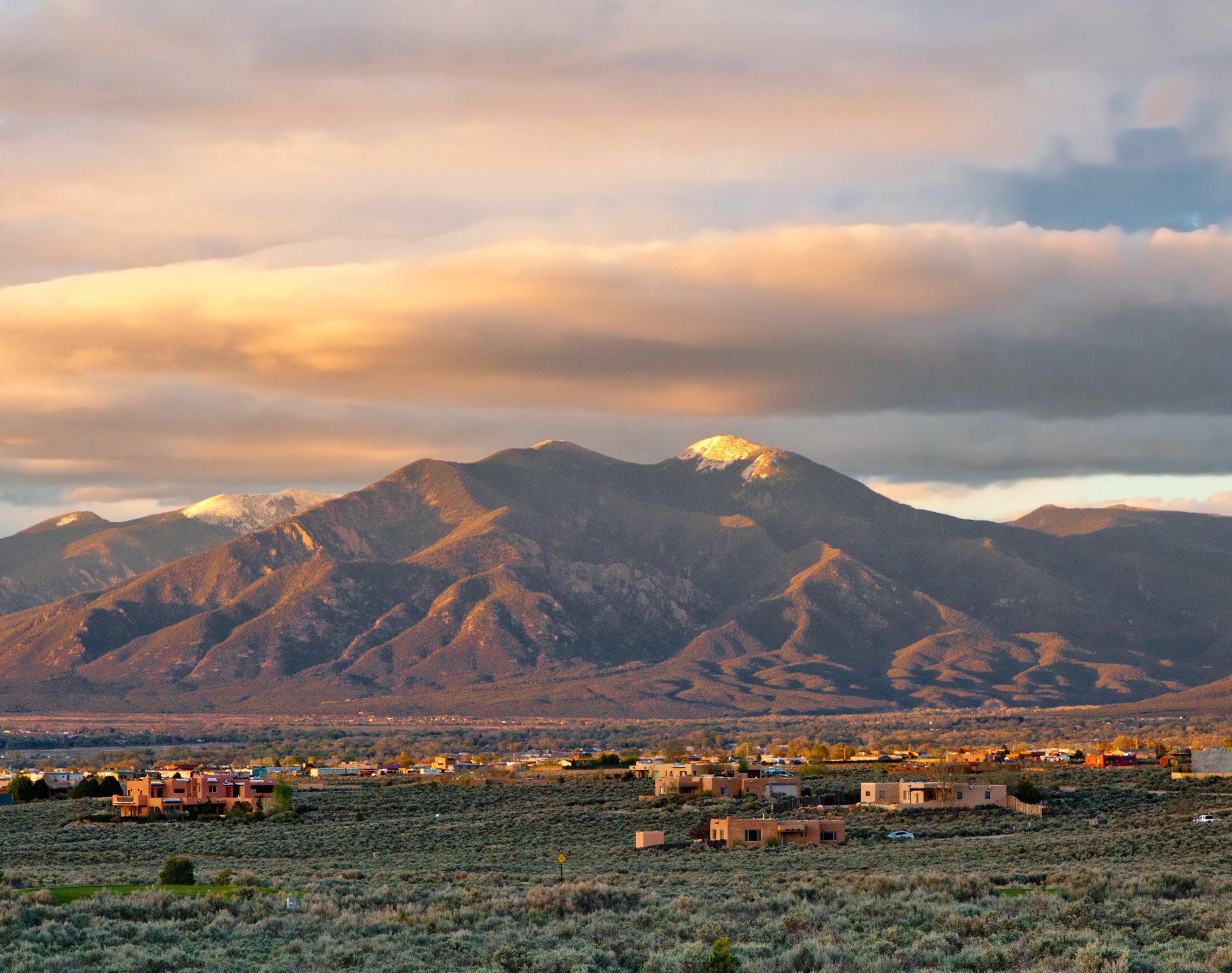 A scenic view of a mountainous landscape with scattered houses at the base, under a sky partly covered with clouds during sunrise or sunset
