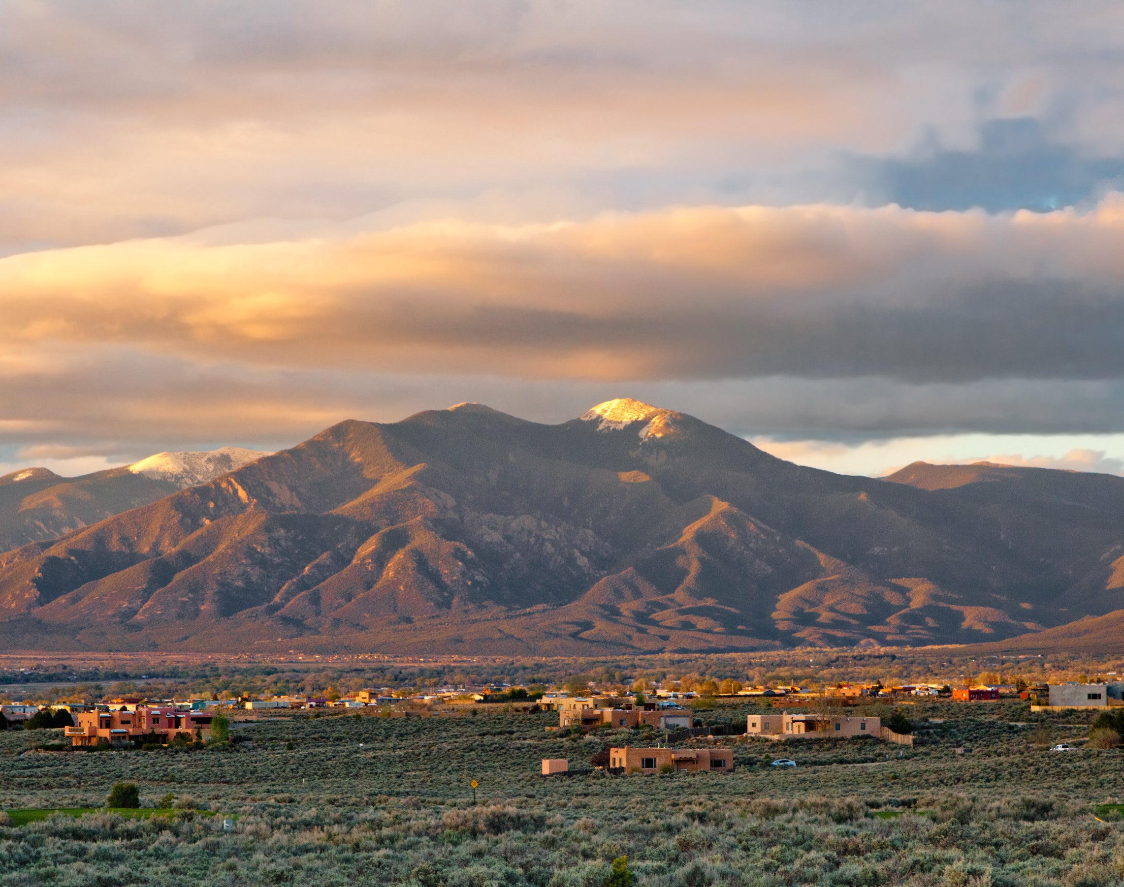 A scenic view of a mountainous landscape with scattered houses at the base, under a sky partly covered with clouds during sunrise or sunset
