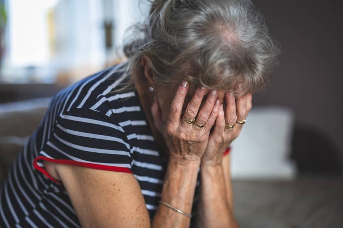 An elderly woman wearing a striped shirt sits on a couch with her head in her hands, appearing distressed