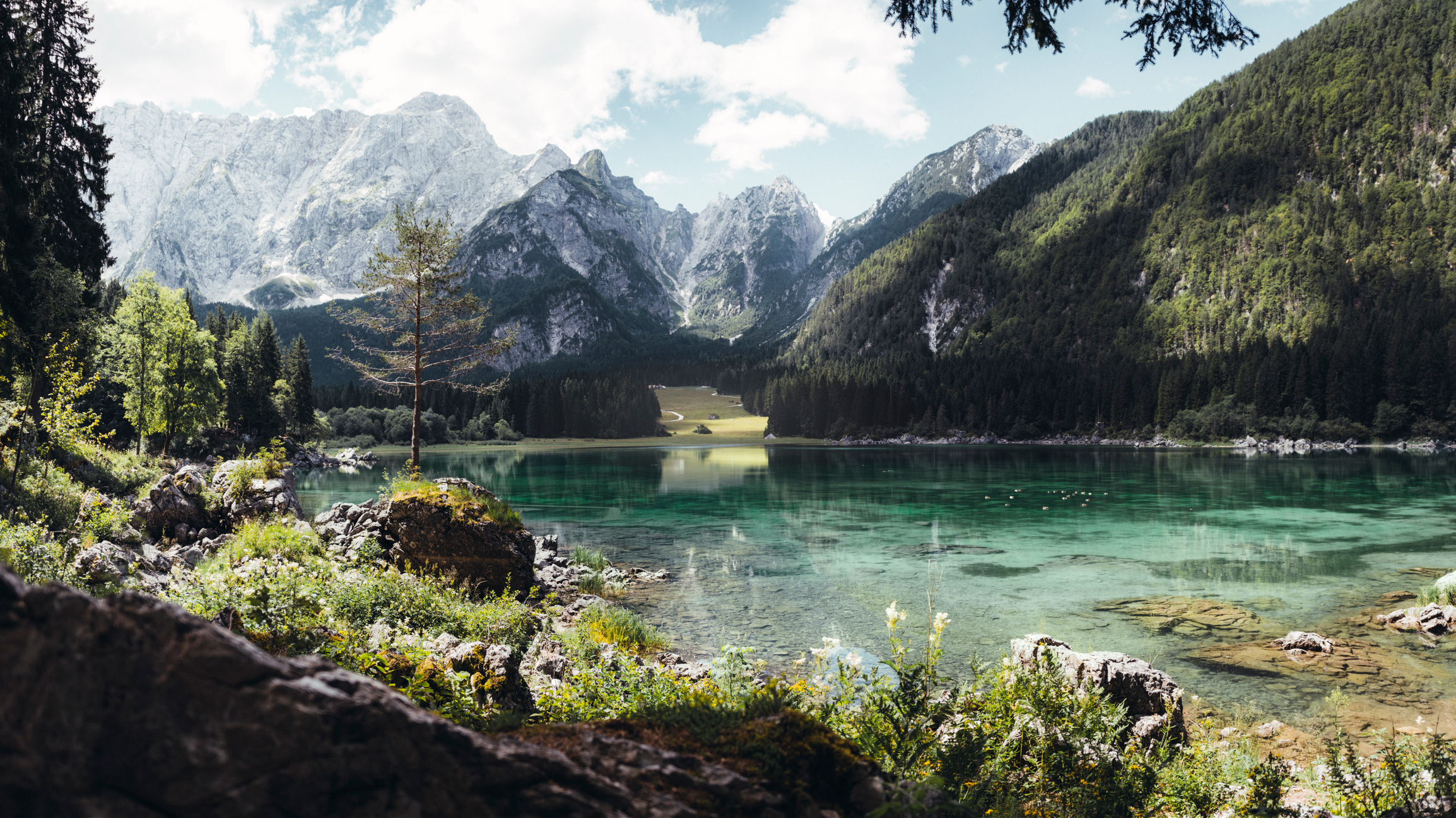 A serene, clear lake surrounded by lush forest and majestic mountains under a partly cloudy sky, forming a peaceful natural landscape
