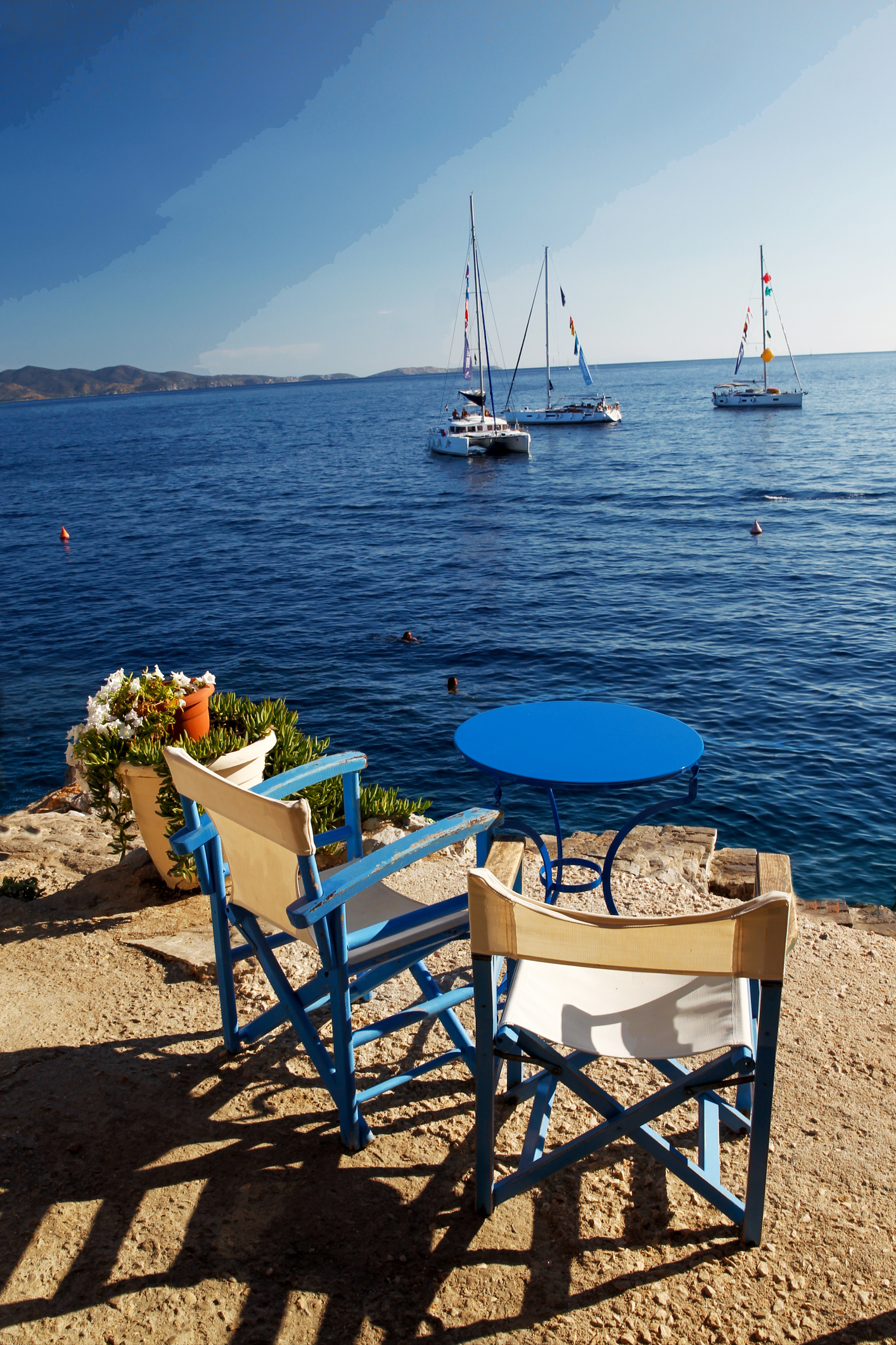 View of three sailboats anchored at sea from a stone patio with two chairs and a table in the foreground. The scene is serene and picturesque