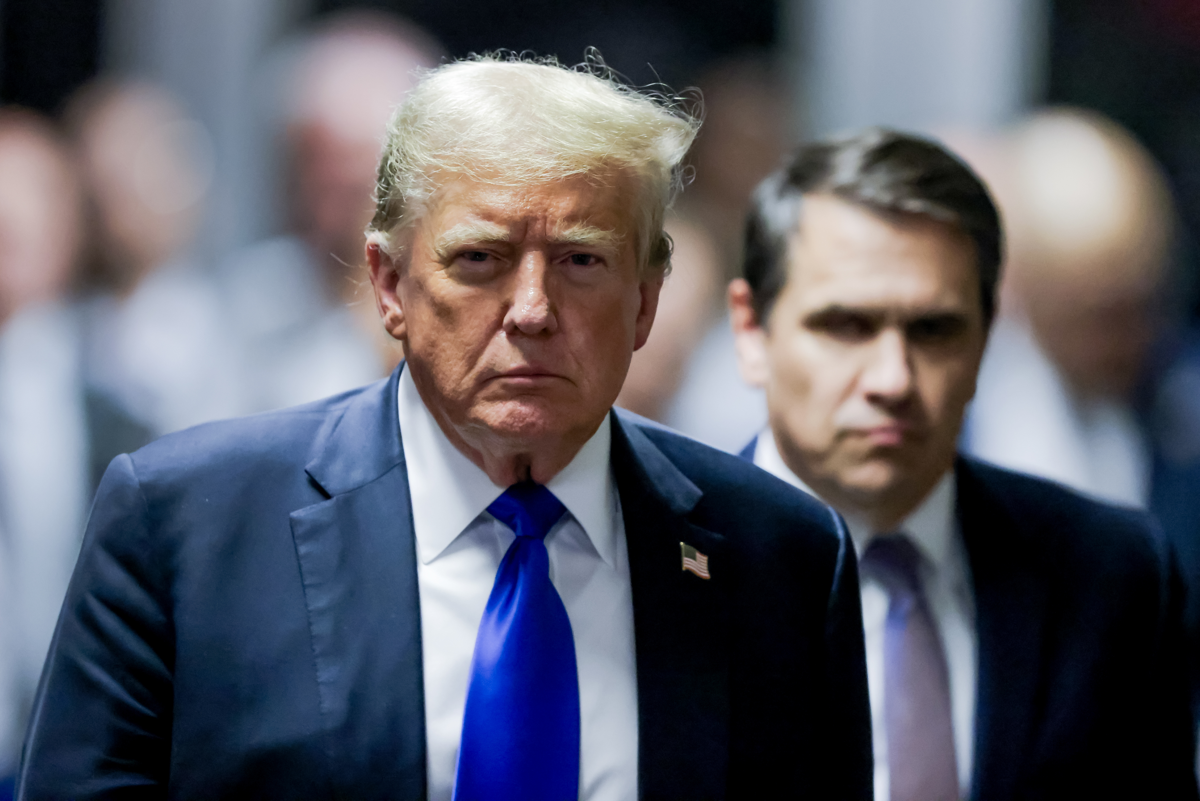 Donald Trump walks with a serious expression alongside an unidentified man. Trump is wearing a dark suit with a blue tie