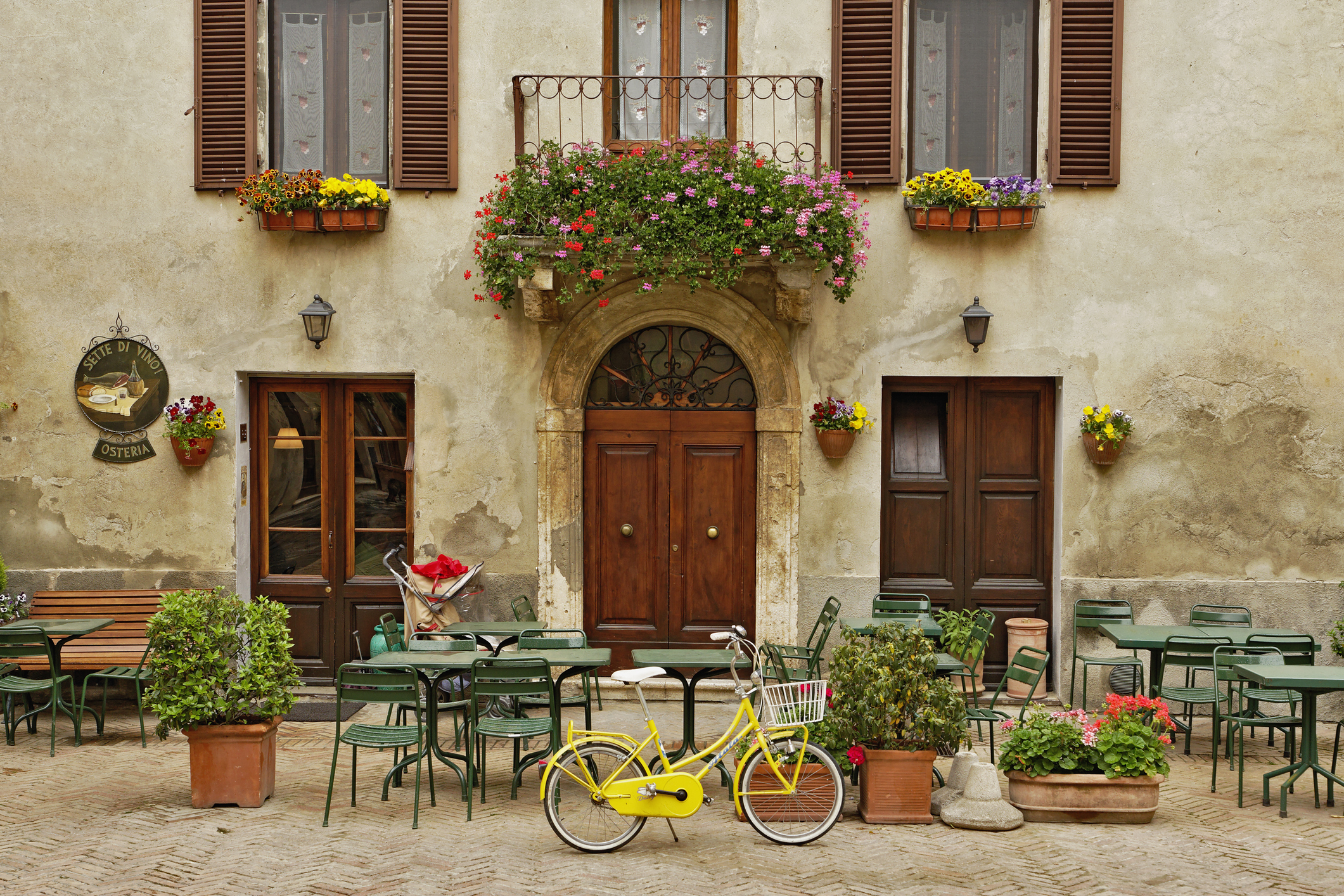 Outdoor café with green tables and chairs, surrounded by potted plants and flowers. A yellow bicycle is parked in front of the café