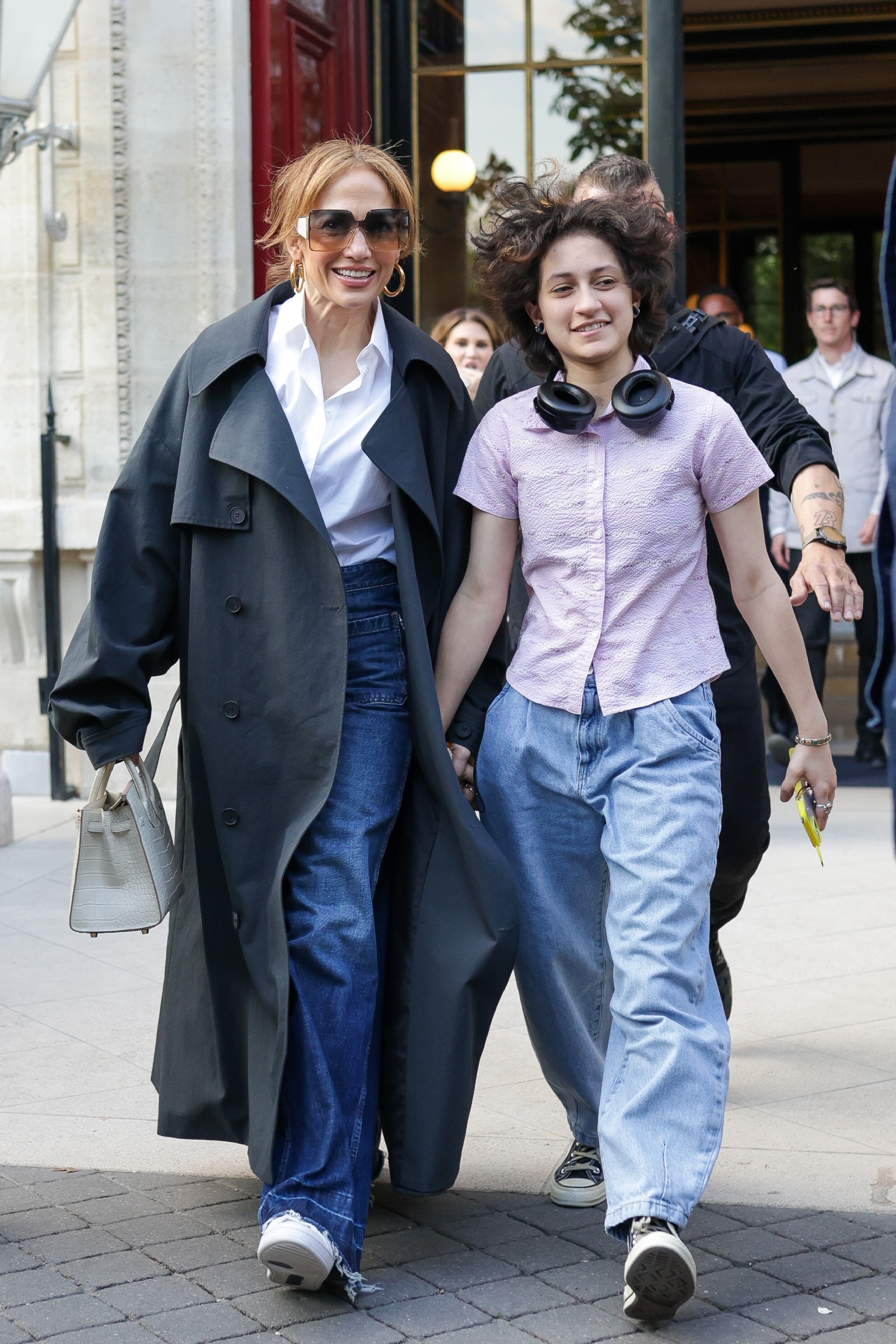 Jennifer Lopez, in a long trench coat, white shirt, and jeans, walks hand-in-hand with her child, Emme Muñiz, sporting a casual shirt, jeans, and headphones