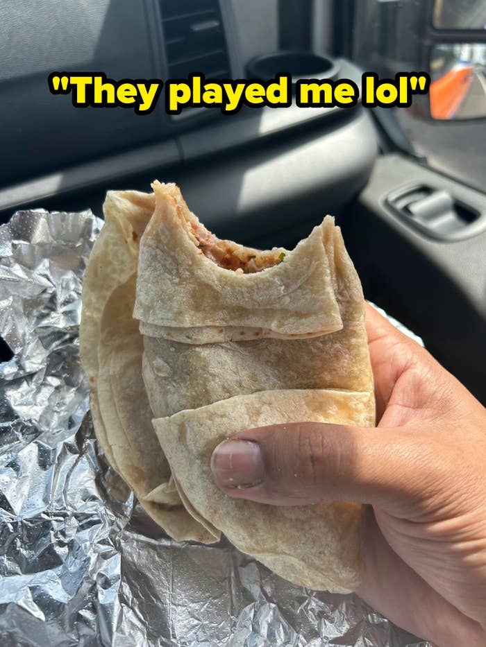Hand holding a close-up of a half-eaten burrito wrapped in foil, inside a car