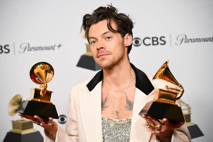 Harry Styles holds two Grammy awards while wearing a sparkling top under a light-colored jacket