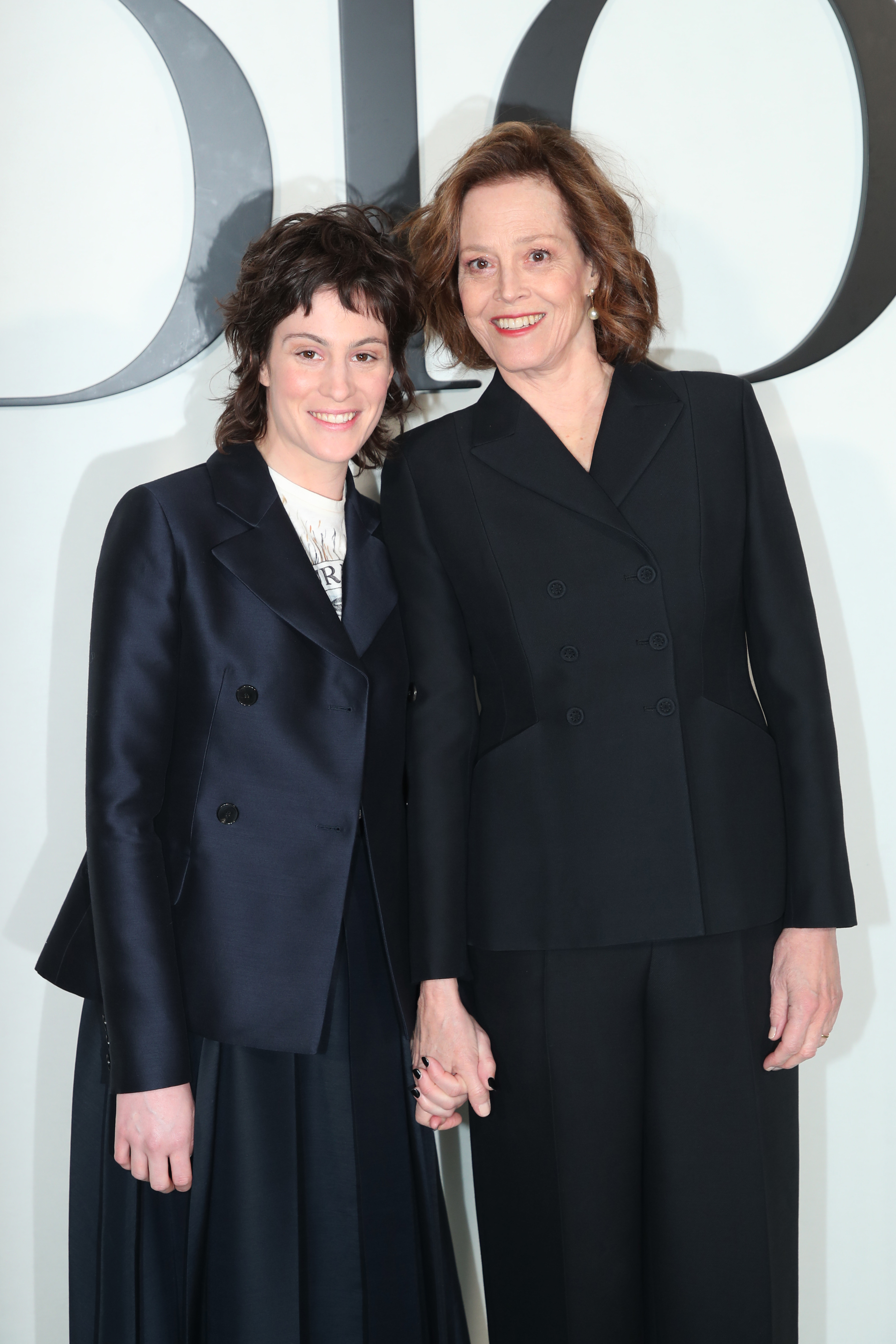Margaret Qualley and Sigourney Weaver smile while holding hands. Both wear dark structured suits at a Dior event
