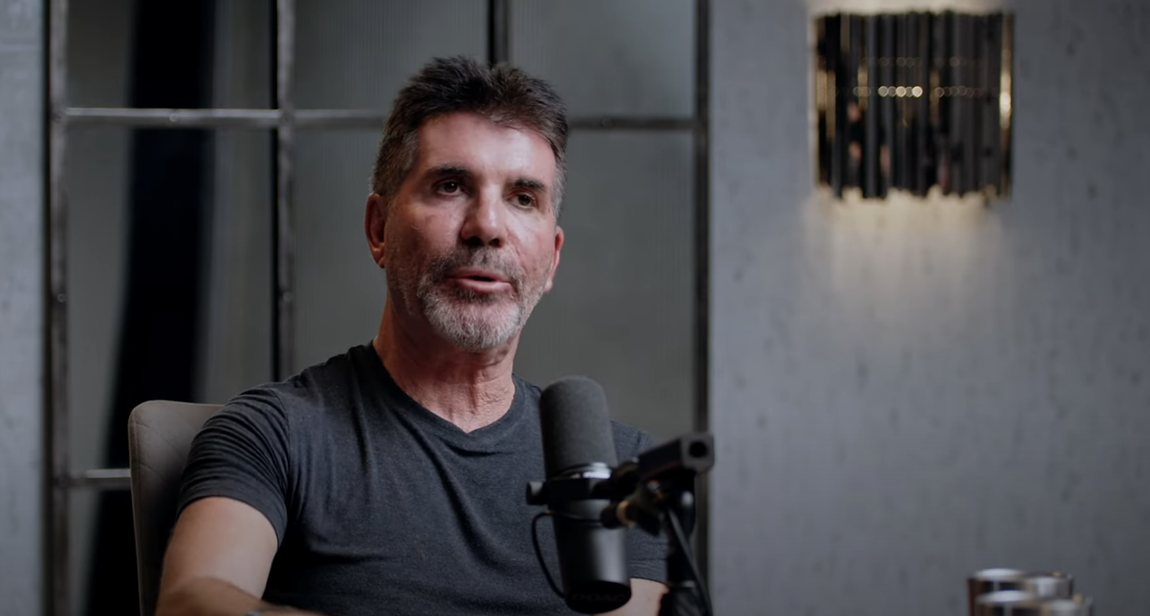 Simon Cowell speaks into a microphone while seated. He wears a casual T-shirt. A window and a wall light fixture are visible in the background