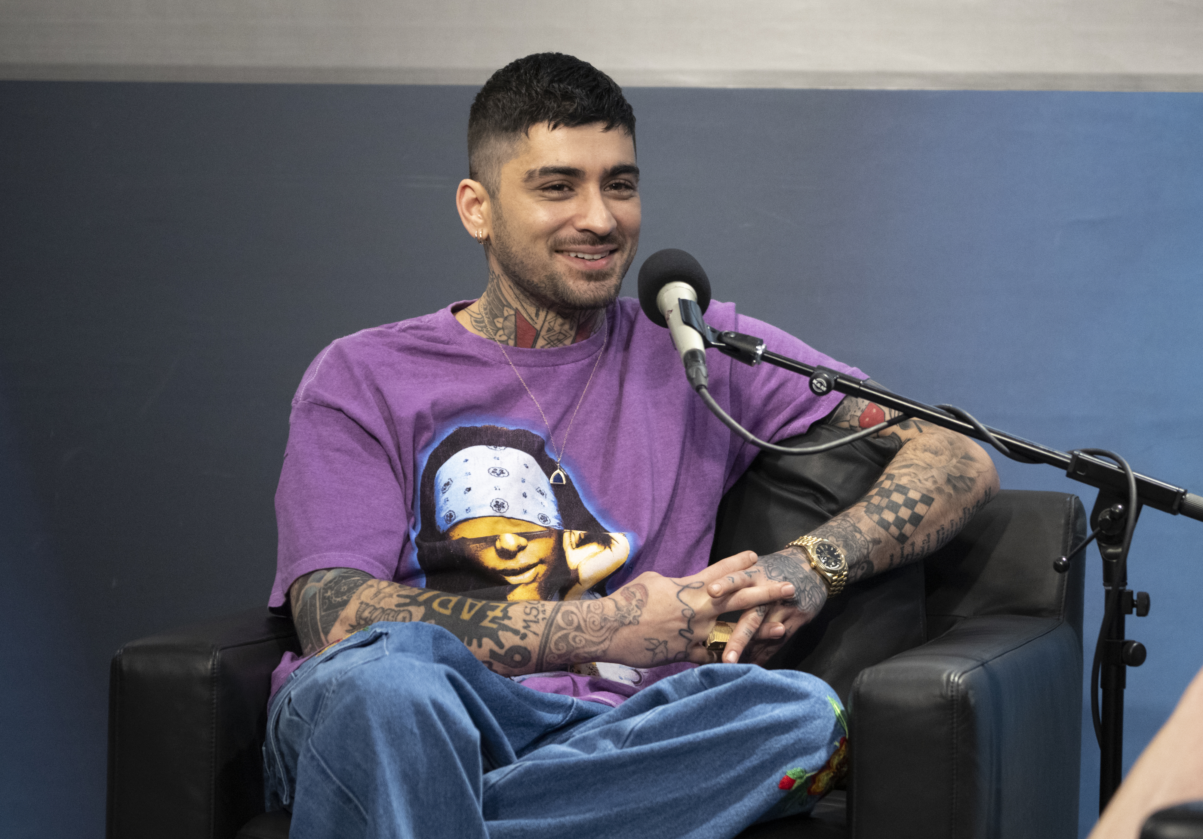 Zayn Malik, wearing a casual outfit with a T-shirt and his tattoos visible, is sitting on a chair while speaking into a microphone during an interview