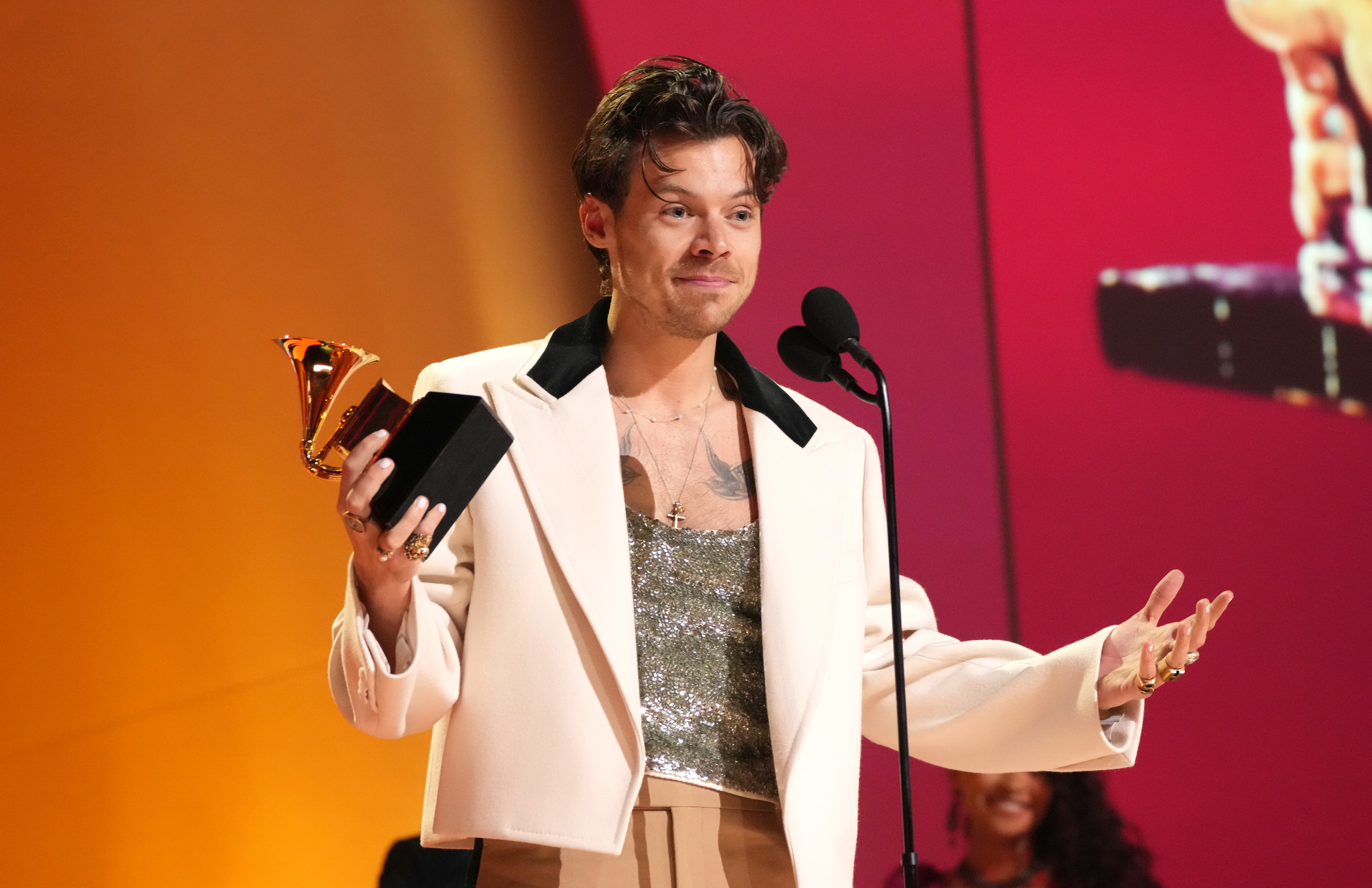 Harry Styles on stage wearing a light jacket and sequin top holding a Grammy award and speaking into a microphone