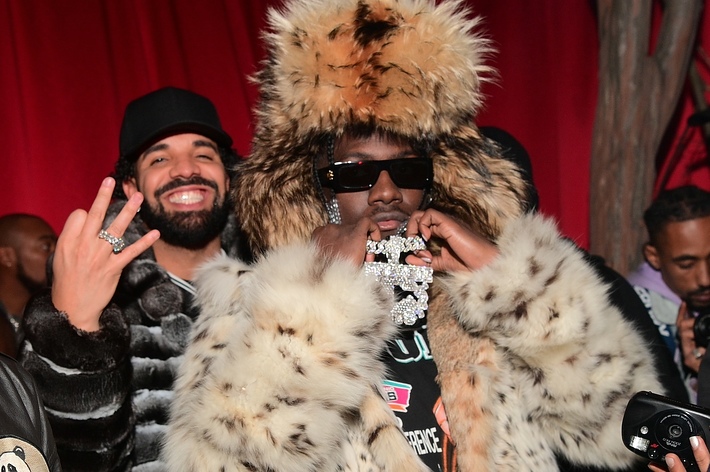 Drake smiles and throws a hand sign while standing behind Lil Yachty, who wears a large fur hat and coat, and holds a diamond-encrusted medallion