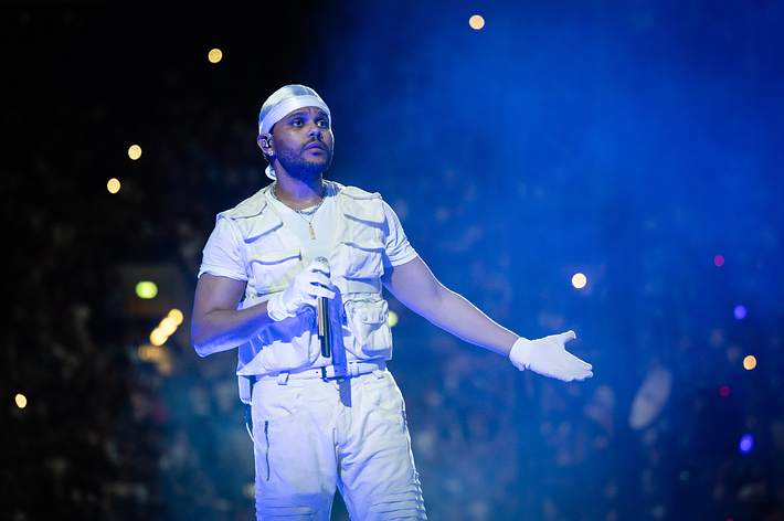 The Weeknd performs on stage wearing a white outfit, including a vest, gloves, and a durag, holding a microphone