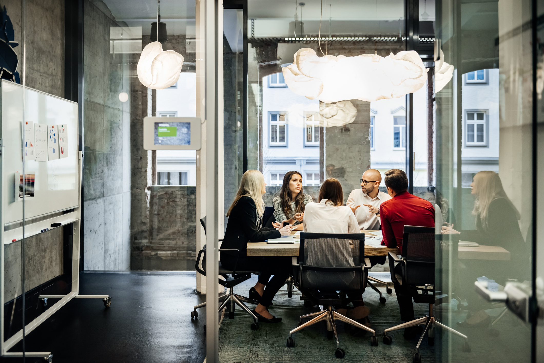 A group of six people are in a modern office meeting room, seated around a table with papers and a whiteboard. The room has large windows and contemporary lighting