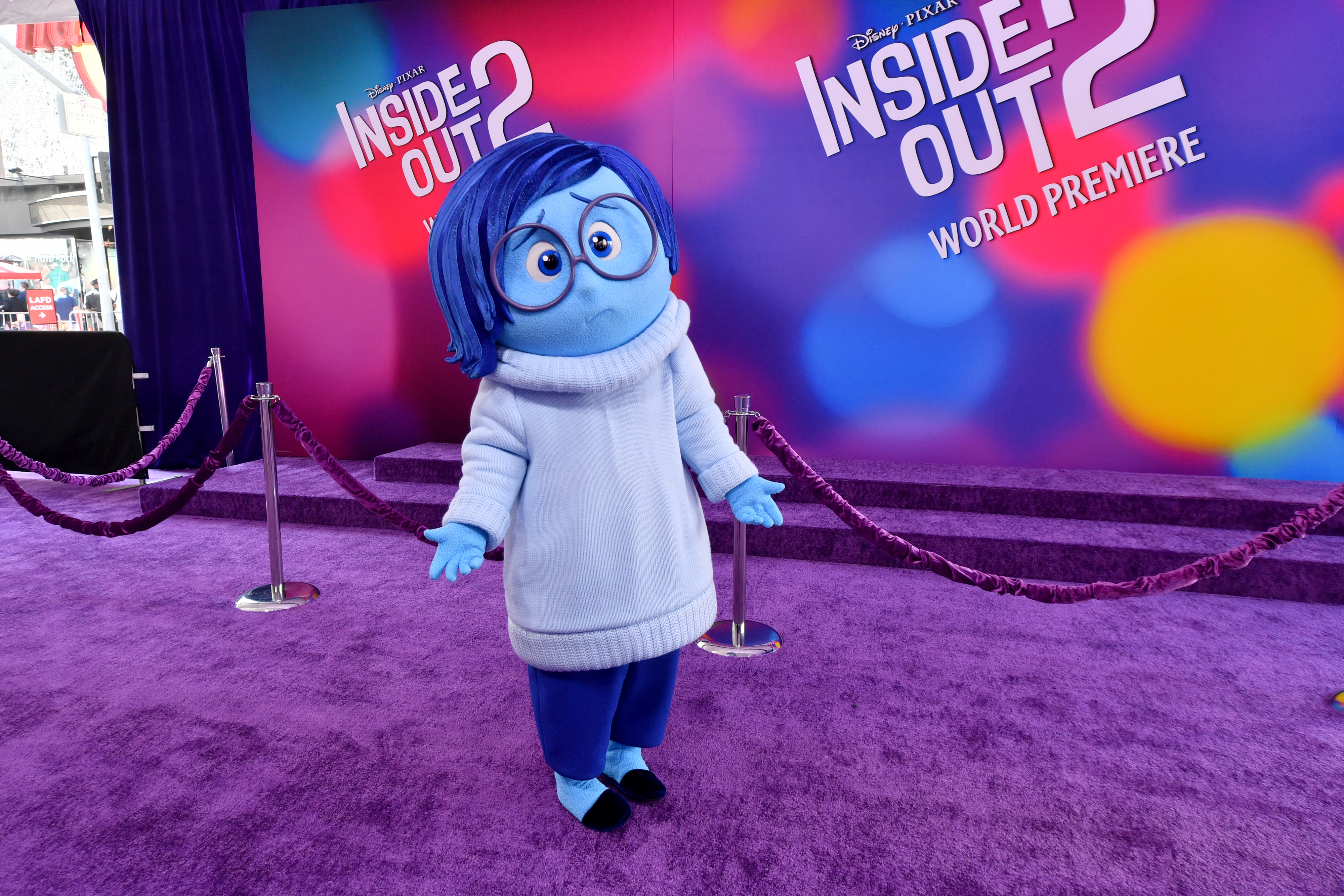 Sadness from Inside Out stands on the purple carpet at the Inside Out 2 world premiere, wearing her signature blue turtleneck and glasses