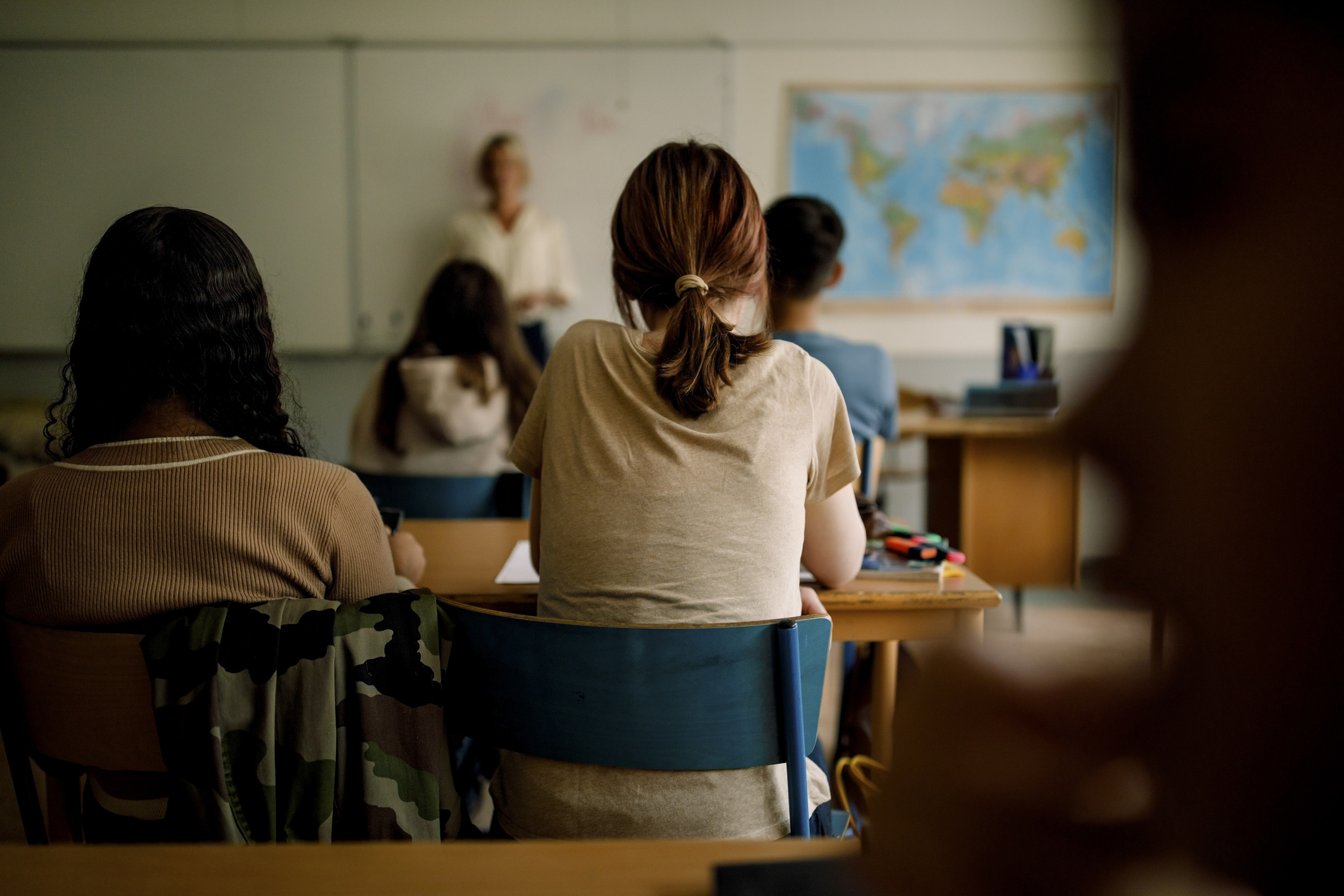 A classroom with students sitting at desks facing a teacher who is standing at the front, next to a whiteboard and map