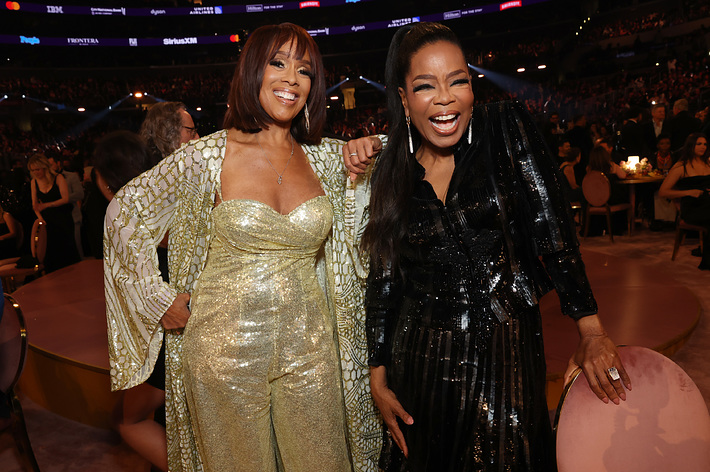 Gayle King and Oprah Winfrey are standing together smiling at an event. Gayle is wearing a shimmering gold outfit, and Oprah is dressed in a sequined black dress