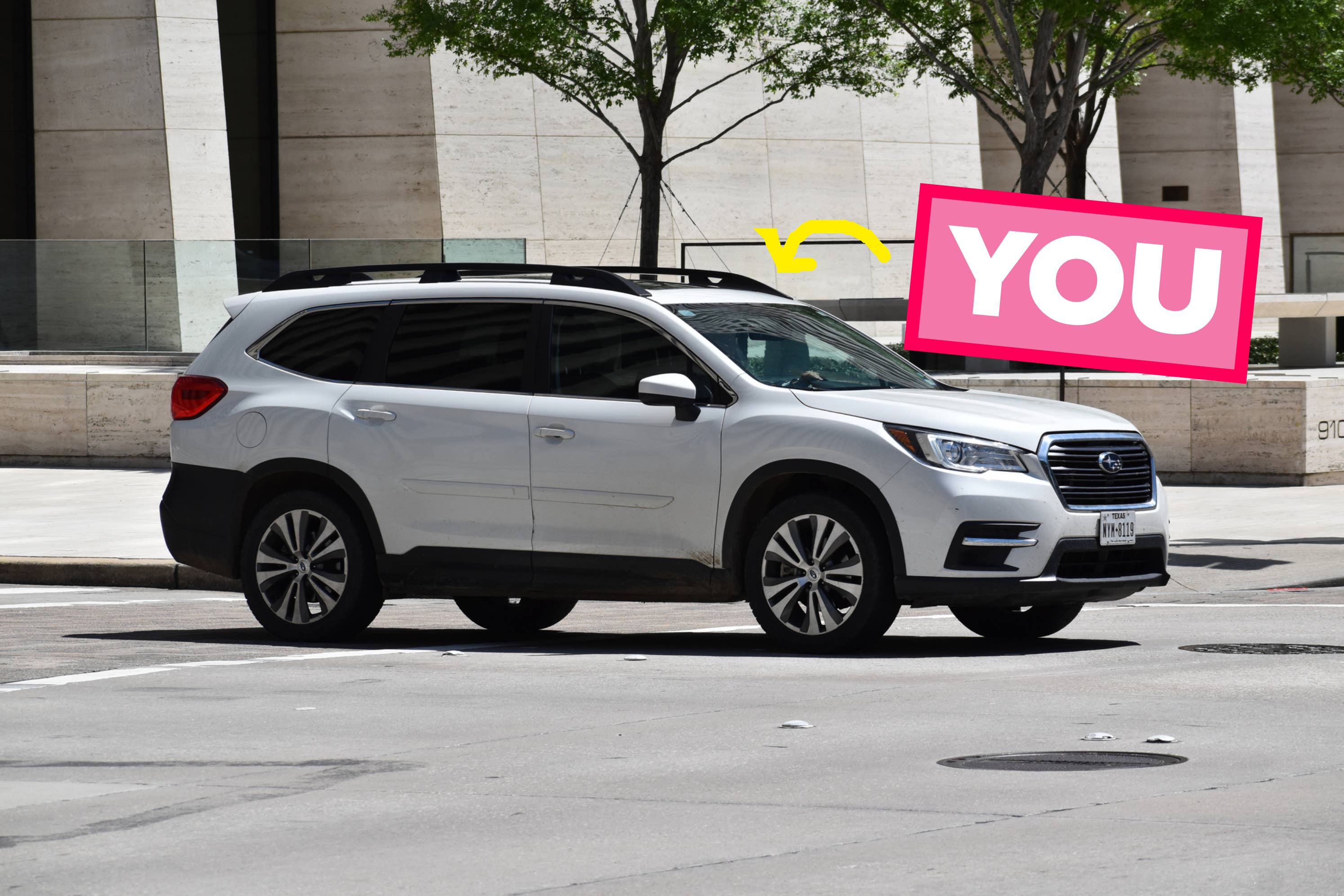 Captioned image of a white SUV on a city street with a pink sign and yellow arrow above pointing to the car, containing the text "YOU"