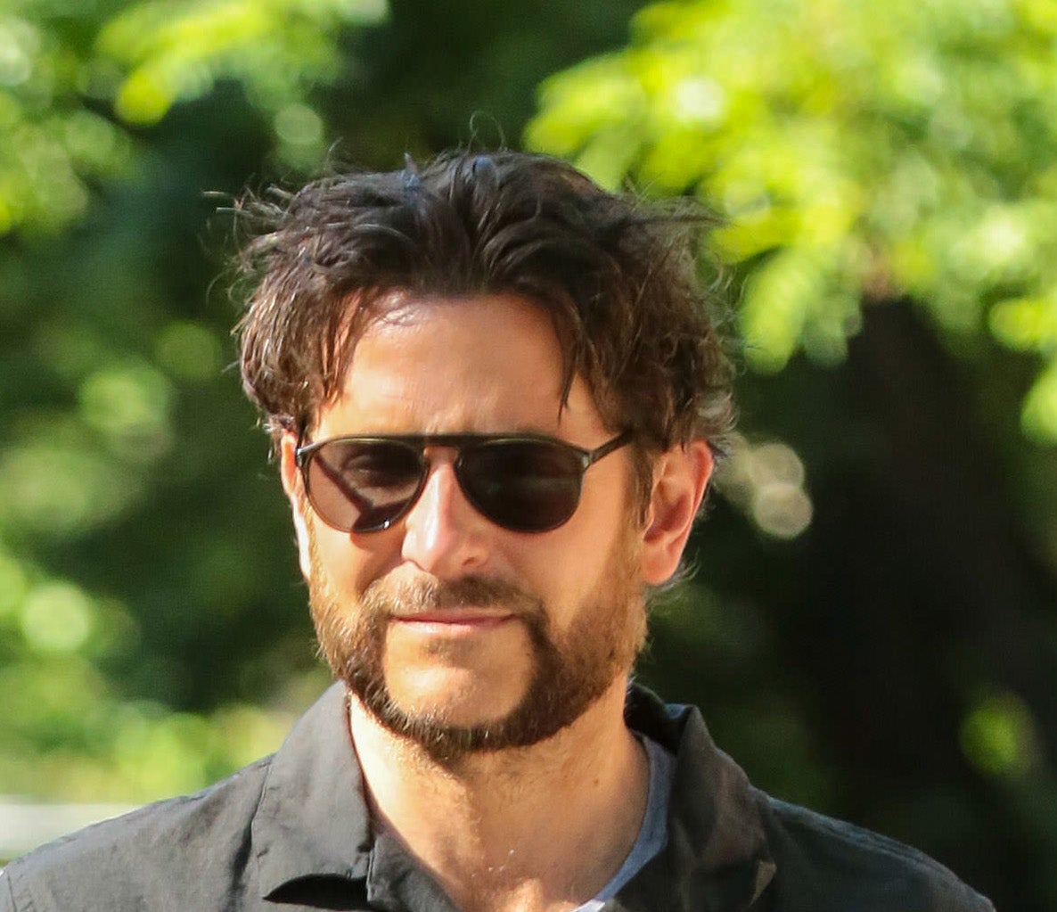 I don't know who this is. A man with sunglasses and a beard is walking outside, wearing a black button-up shirt with white patterns and blue jeans, holding a phone