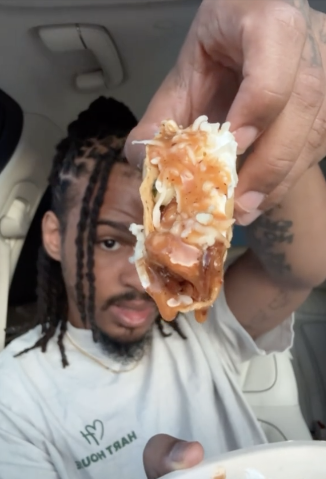 Keith Lee holding a taco filled with cheese, meat, and sauce up to the camera. He appears to be in a vehicle