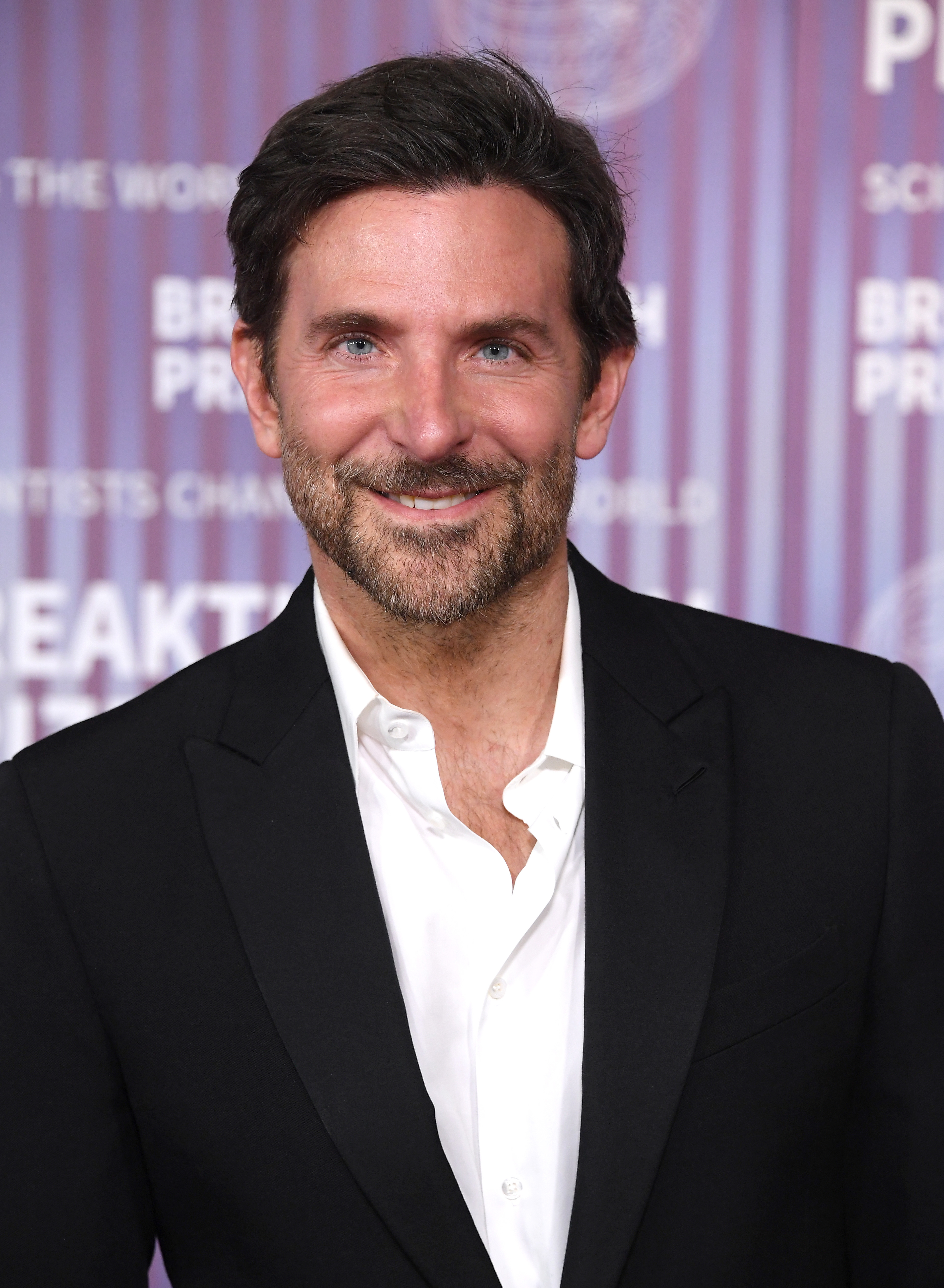 Bradley Cooper smiling, wearing a black suit jacket over a white dress shirt, at a red carpet event. Background features blurred text and design