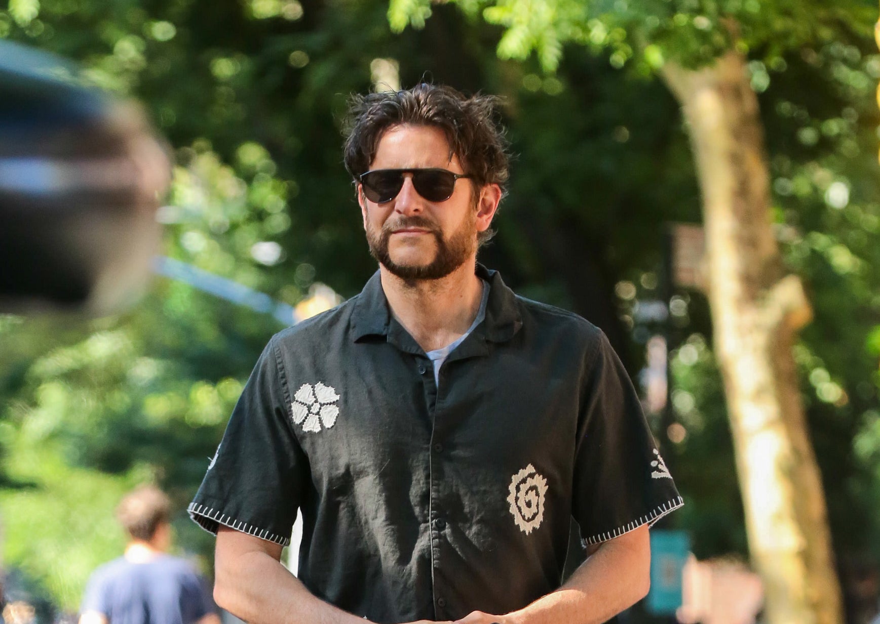 Michael Zegen walks down a city street wearing sunglasses, a black patterned shirt, and jeans