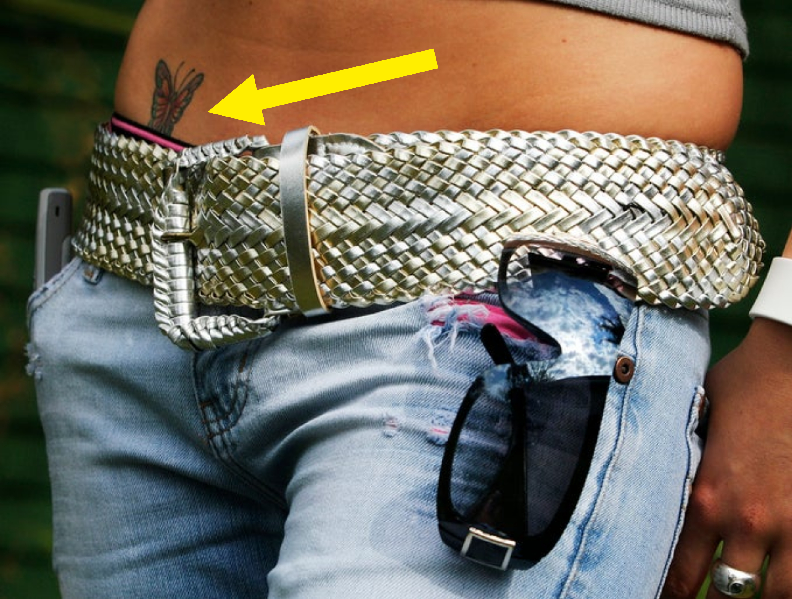 A close-up of a person's waist wearing blue jeans, a thick woven belt, and sunglasses hanging from the jeans pocket. A butterfly tattoo is visible above the jeans