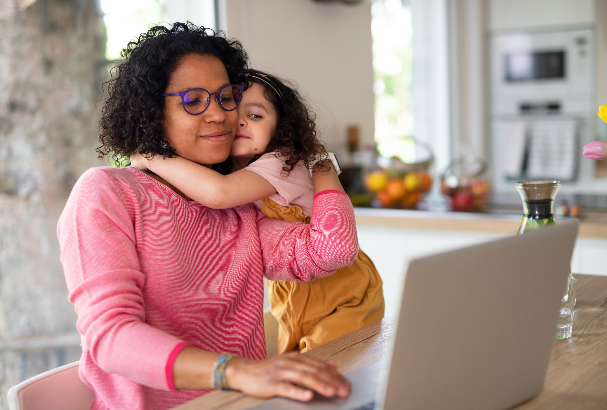 A mother in glasses, wearing a pink sweater, works on a laptop while her young daughter hugs her from behind in a cozy kitchen setting
