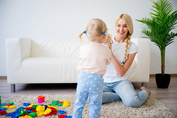 A woman with a long braid, wearing a white shirt and jeans, sits on the floor and claps hands with a toddler in polka dot pants playing with toys