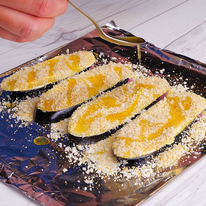 アルミホイルに並んだ4枚のパン粉をまぶしたナスに油をかける手。