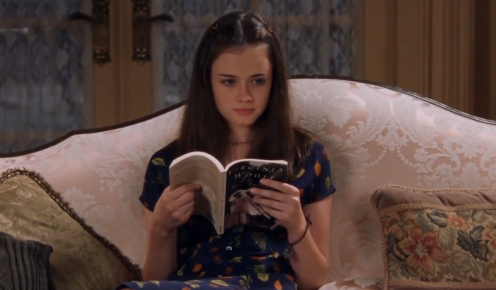 A young girl, Alexis Bledel, sits on a sofa reading a book. She wears a patterned dress and appears focused on her reading