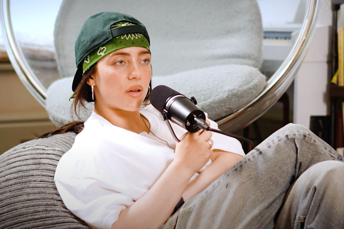 Billie Eilish is lounging in a chair, wearing a casual white shirt and gray pants, with a green bandana and hat, holding a microphone in a recorded interview