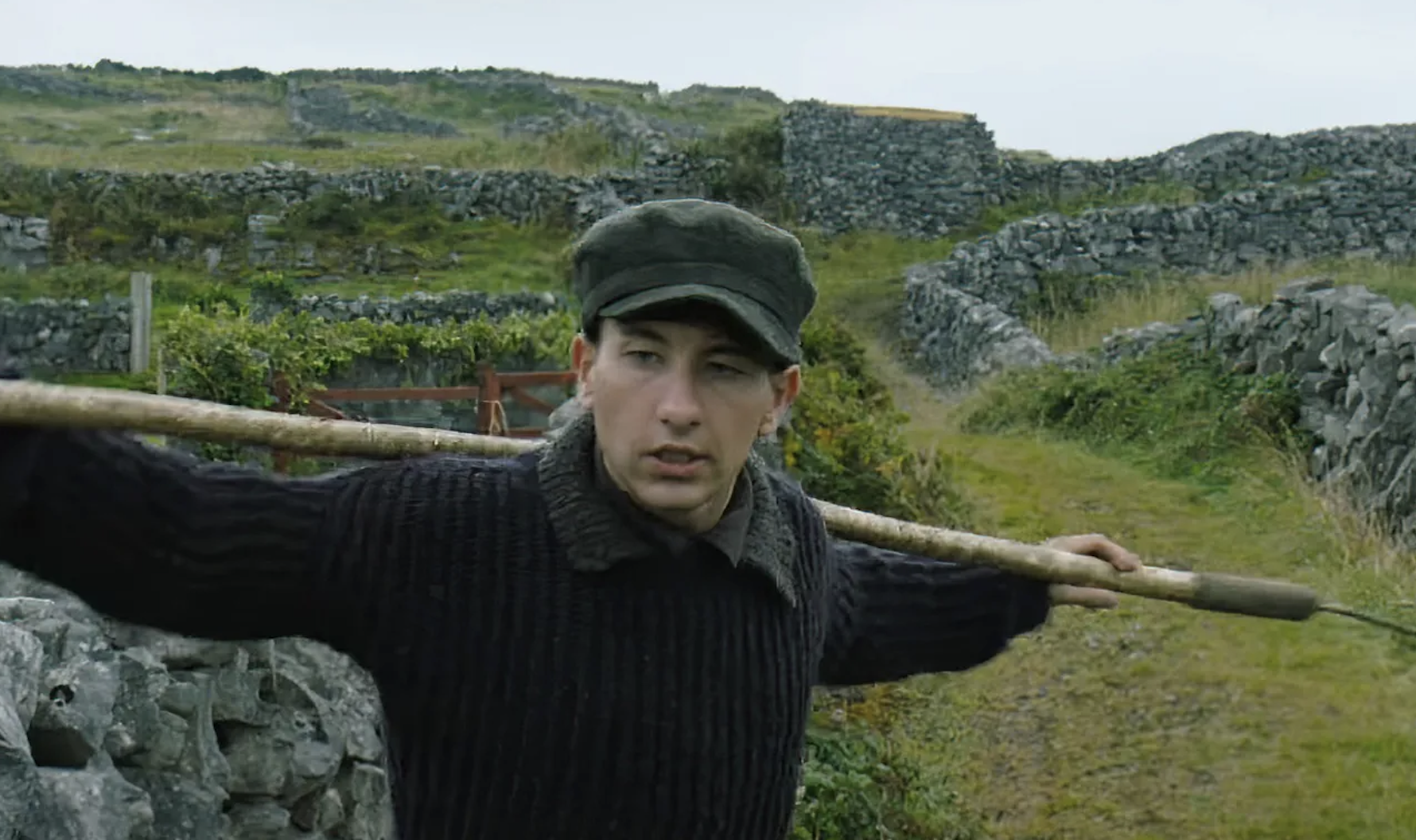 Barry Keoghan stands outdoors on a rocky path, carrying a wooden pole over his shoulder, wearing a knit sweater and a flat cap