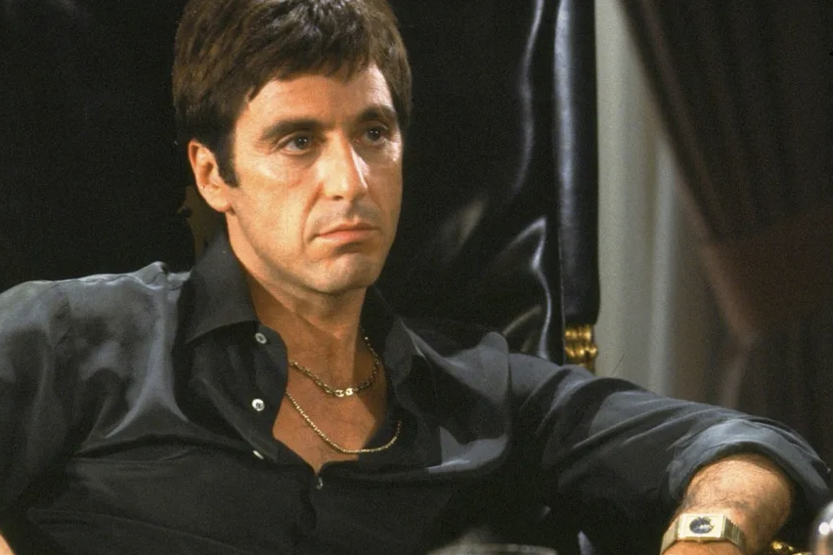 Al Pacino as Tony Montana from the movie Scarface, sitting in a chair, wearing a dark shirt and gold chains