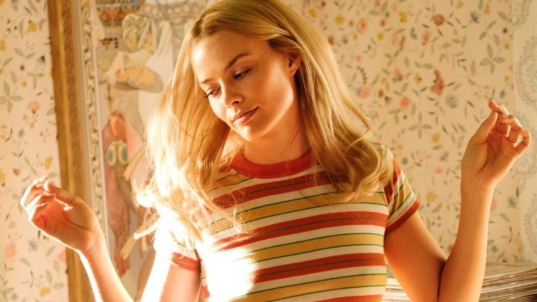 Margot Robbie, wearing a striped short-sleeve top, stands with her arms slightly raised in front of a floral patterned background