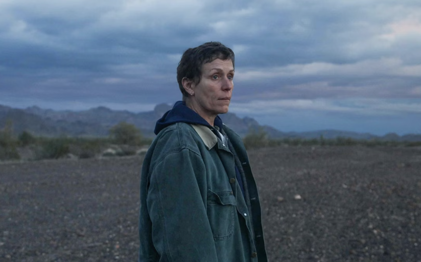 Frances McDormand stands outdoors in a barren landscape, looking into the distance, with mountains and a cloudy sky in the background