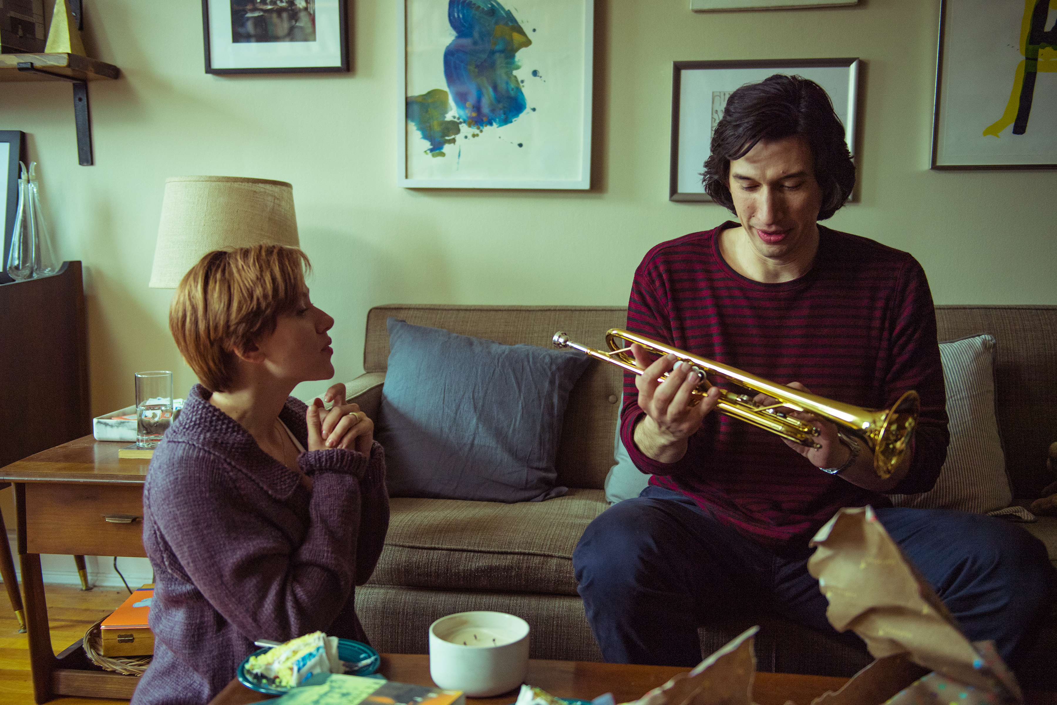 Scarlett Johansson and Adam Driver sit in a living room. Scarlett is looking at Adam, who is holding a trumpet and seems to be talking. There is a bowl on the table