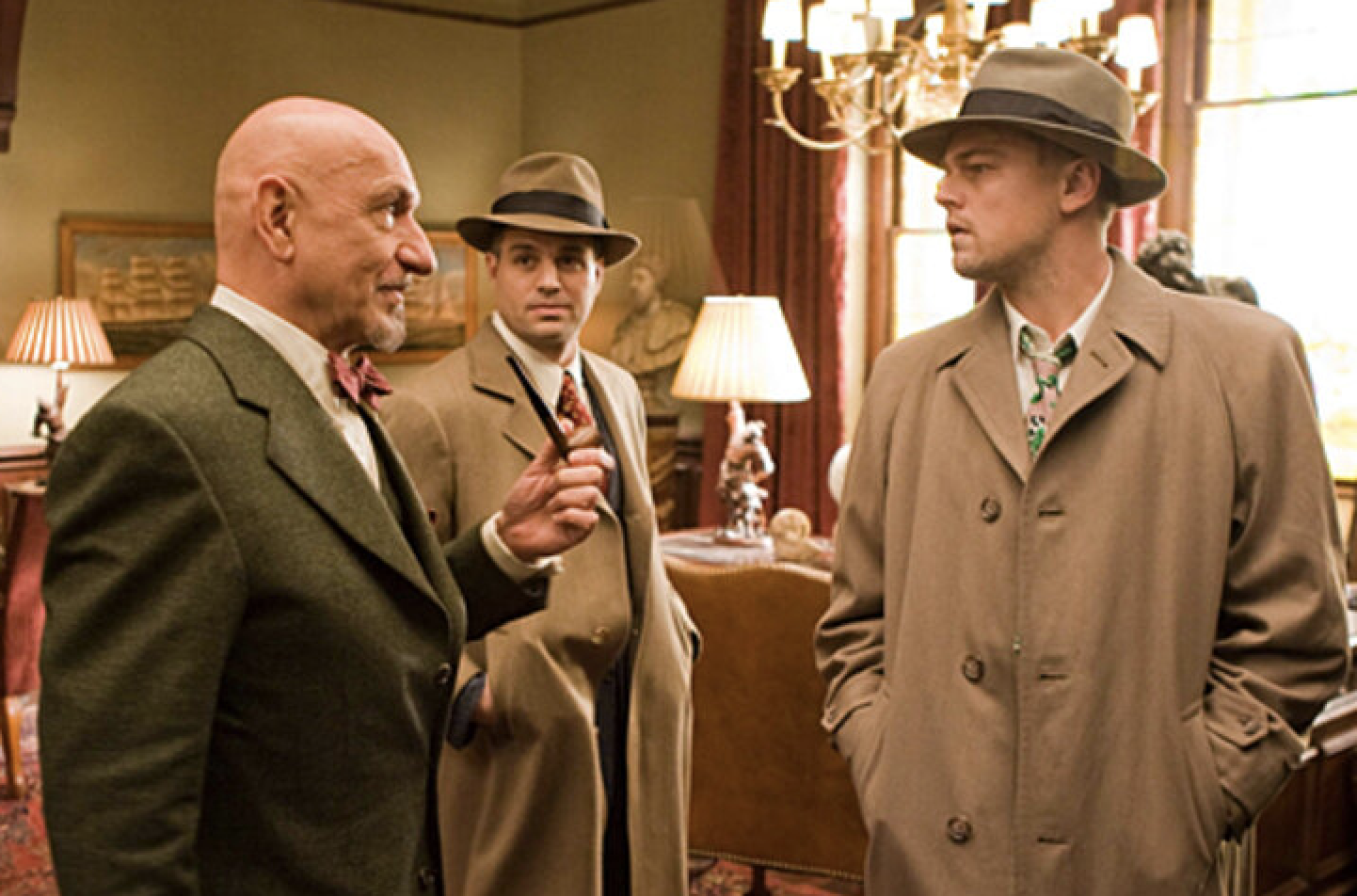 Ben Kingsley, Mark Ruffalo, and Leonardo DiCaprio in a vintage room. Kingsley in a bowtie, Ruffalo in a suit and fedora, DiCaprio in a trench coat and fedora