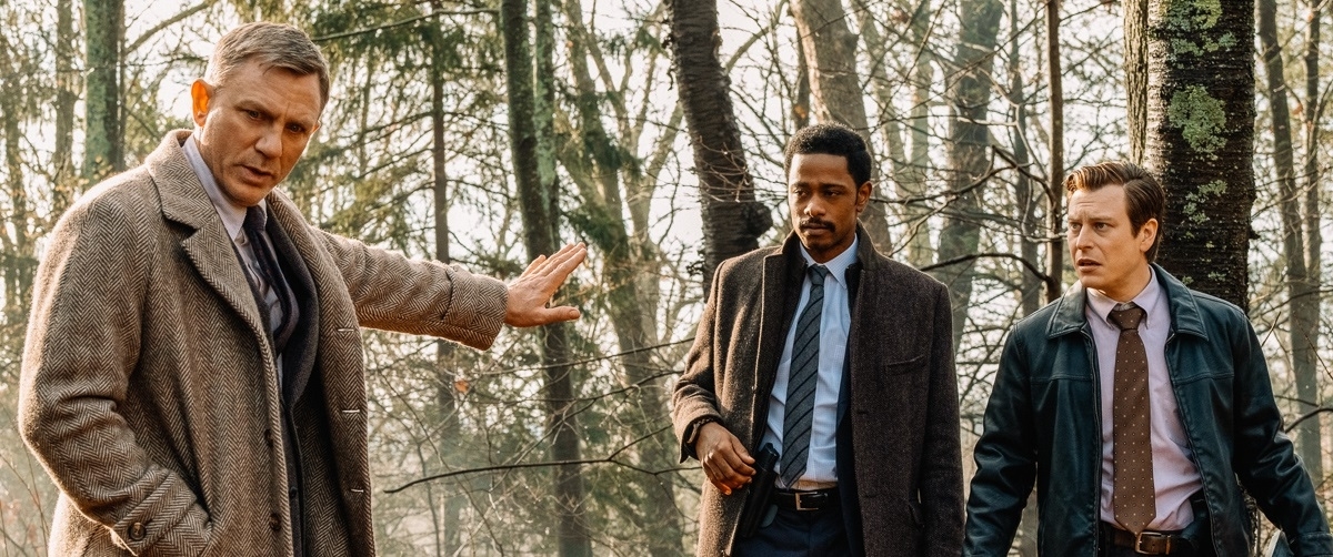 Daniel Craig, Lakeith Stanfield, and Noah Segan in a forest scene, dressed in coats and ties, appear to be discussing something serious