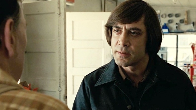 Javier Bardem, dressed in a denim jacket, stands in a room facing to the left, having a serious conversation with a person in a plaid shirt who is partially visible